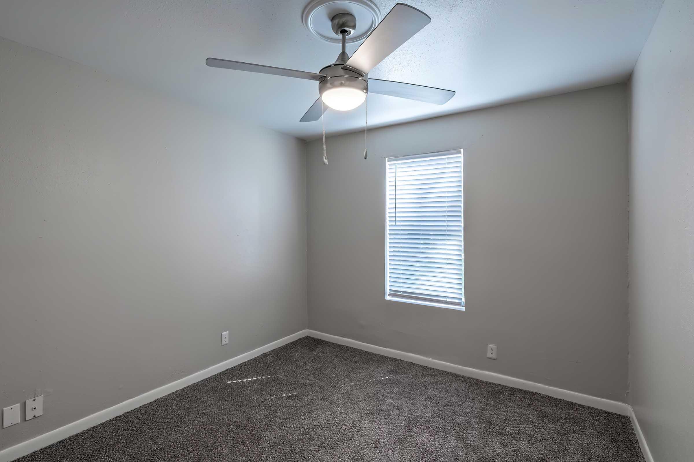A small, empty room with light gray walls and a ceiling fan. A window with white blinds allows natural light to enter, creating a bright atmosphere. The floor is covered in gray carpet, and there are a few electrical outlets visible on the walls. The room appears clean and uncluttered.