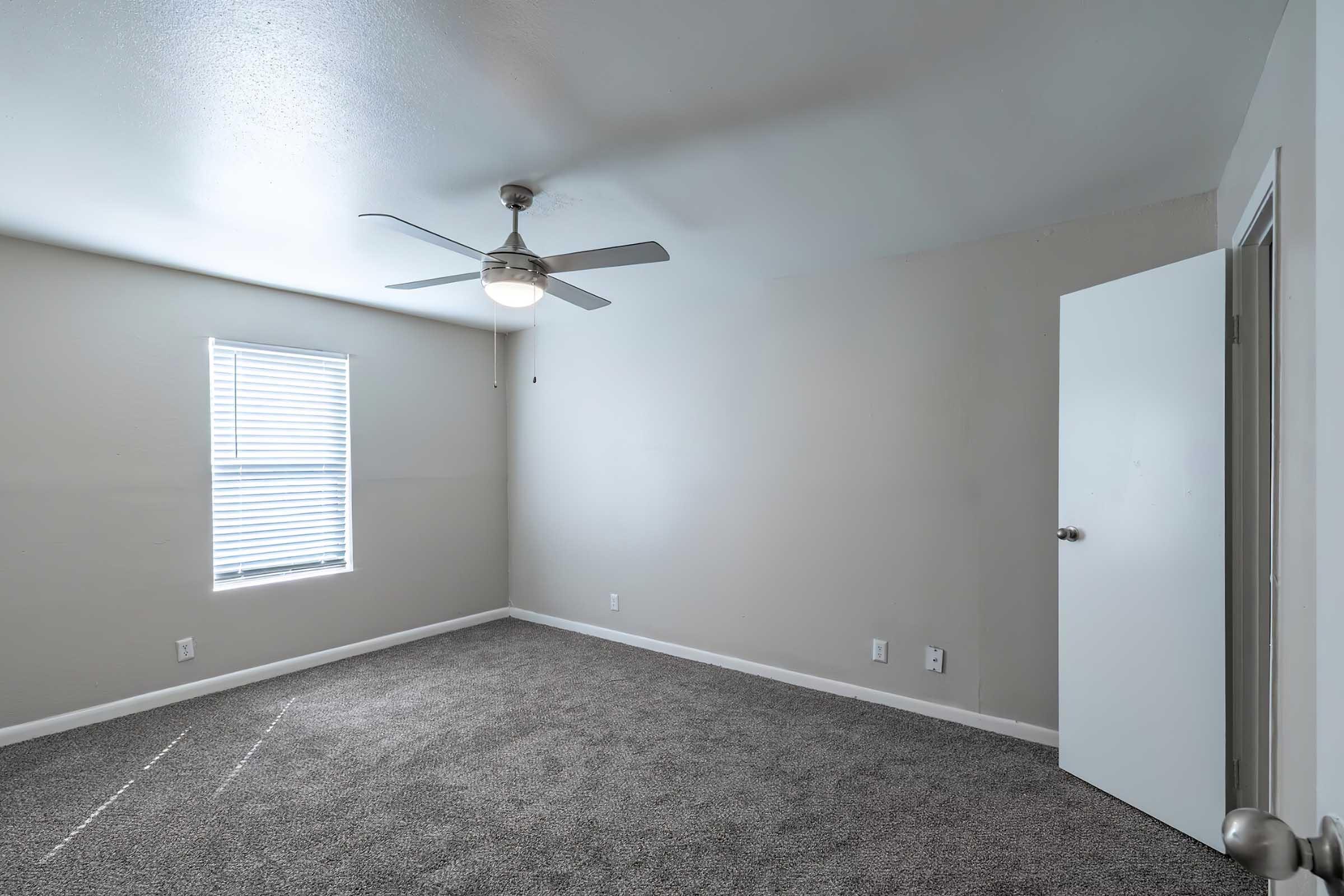 A spacious, empty room with light gray walls and plush carpet. A ceiling fan is overhead, and natural light streams in through a window with blinds. A closed white door leads to another space, indicating a neat and modern aesthetic.