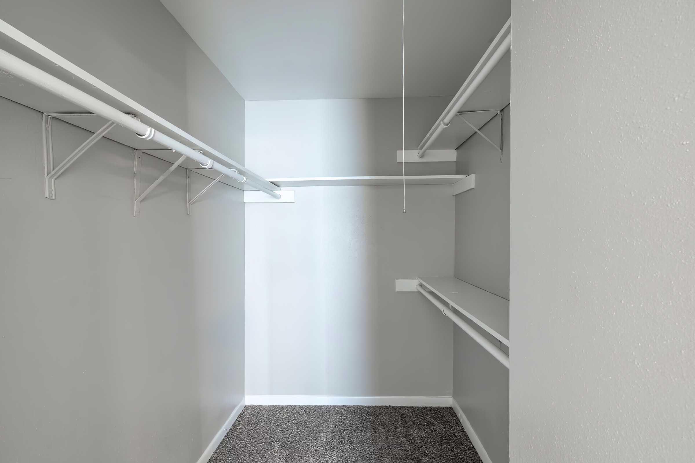 Empty closet with gray walls and carpeted flooring. It features white shelving and hanging rods on opposite walls, with ample space for storage. The lighting creates a bright appearance, emphasizing the open and uncluttered layout.