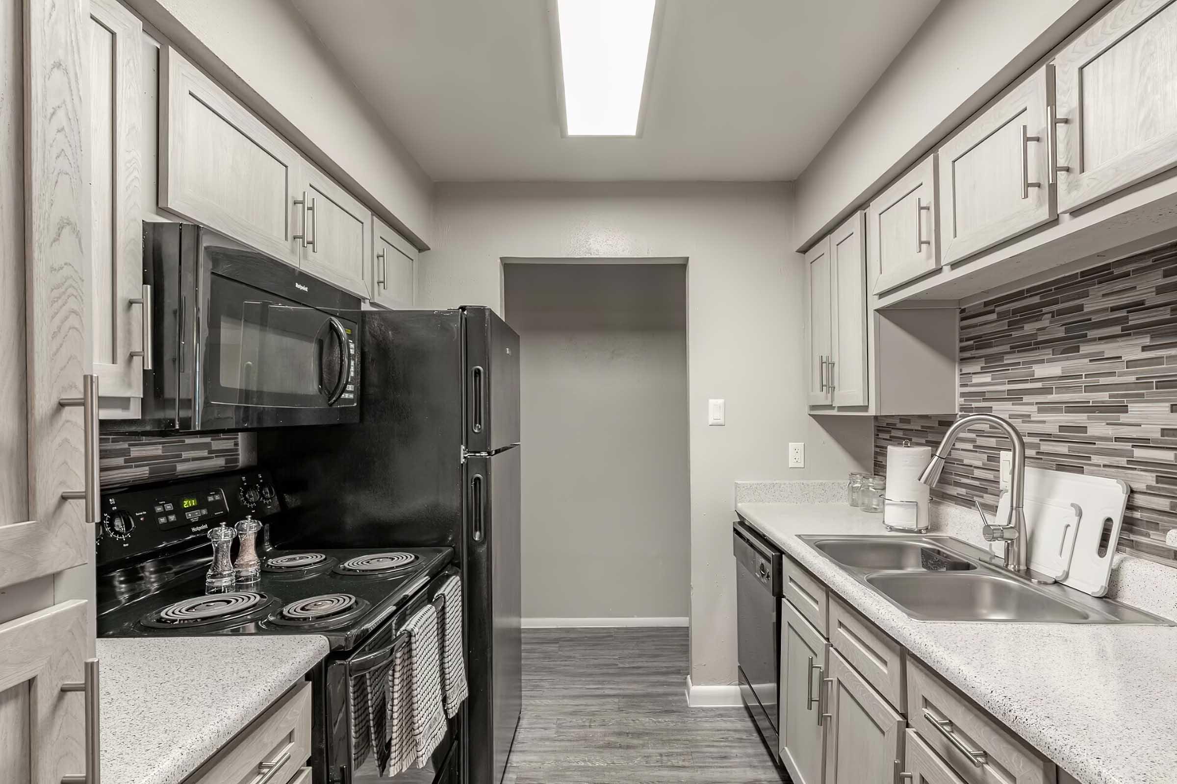 A modern kitchen featuring light wood cabinetry, a black refrigerator, and a black stove. The countertop is light with a textured backsplash in shades of gray. There is a sink and window with minimal decor, and the room is well-lit with overhead fluorescent lighting.
