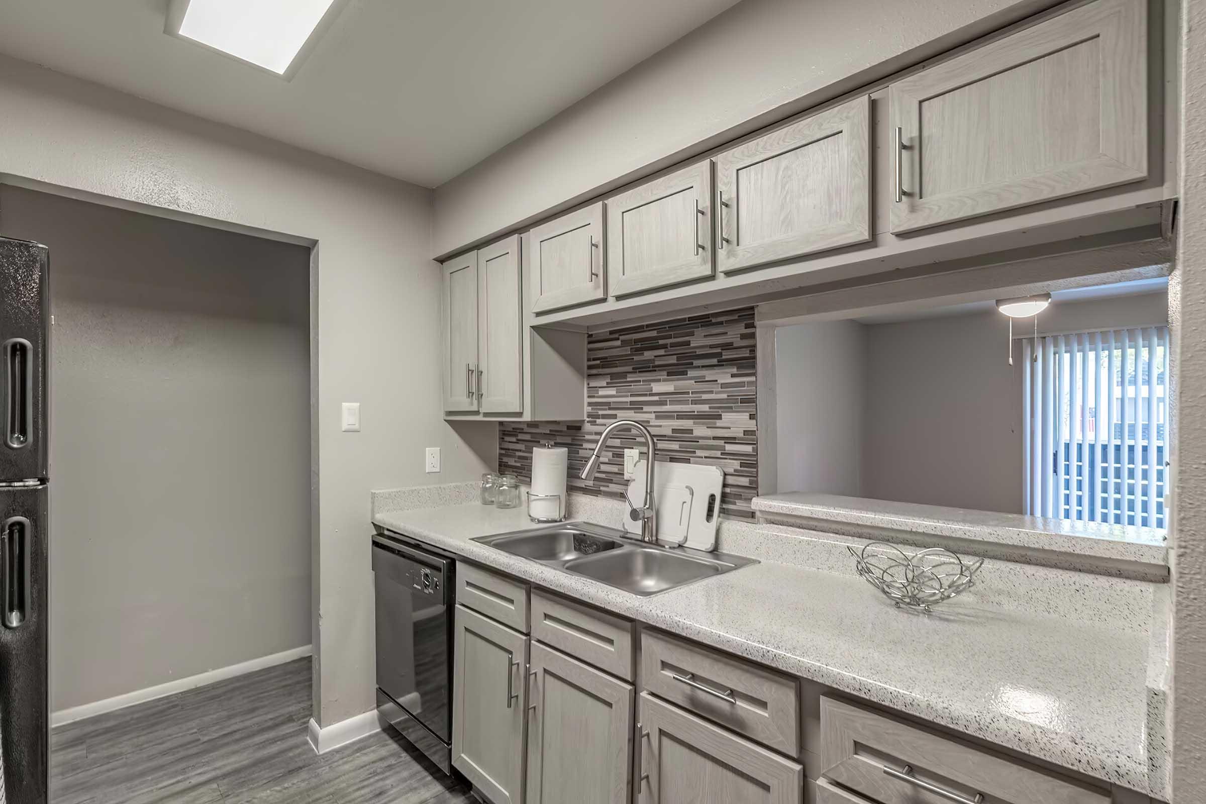 Modern kitchen with grey cabinetry, a double sink, and a dishwasher. The countertop is a light color with a speckled finish. A tiled backsplash features a mix of grey tones. The kitchen opens to a living area with a window covered by vertical blinds, providing a bright and spacious feel.
