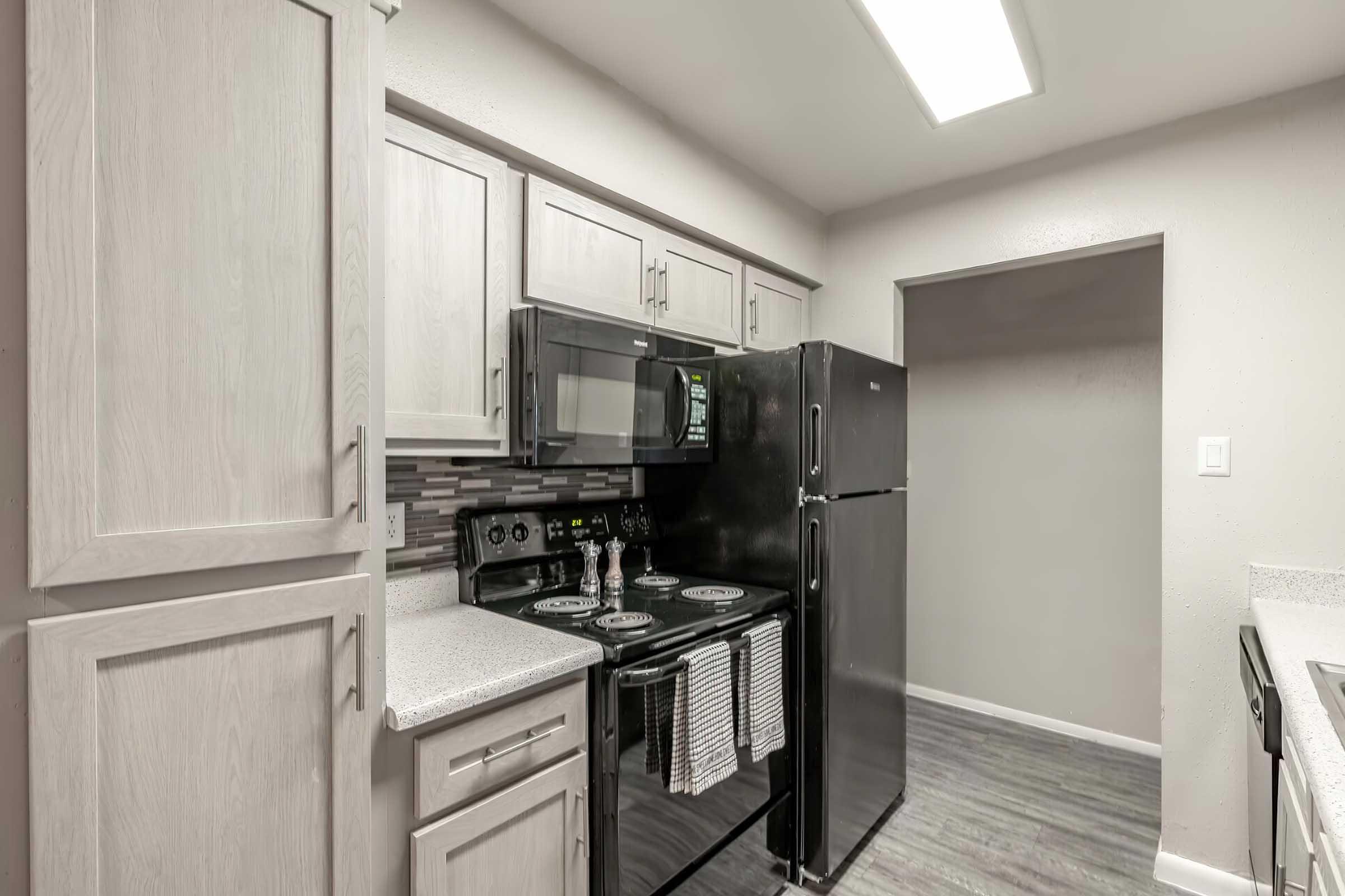 Modern kitchen featuring light wood cabinets, a black stove and microwave, and a black refrigerator. Countertops are light-colored, and there are decorative items on the stove. The space is well-lit with a bright ceiling light, and there is an open doorway leading to another area.