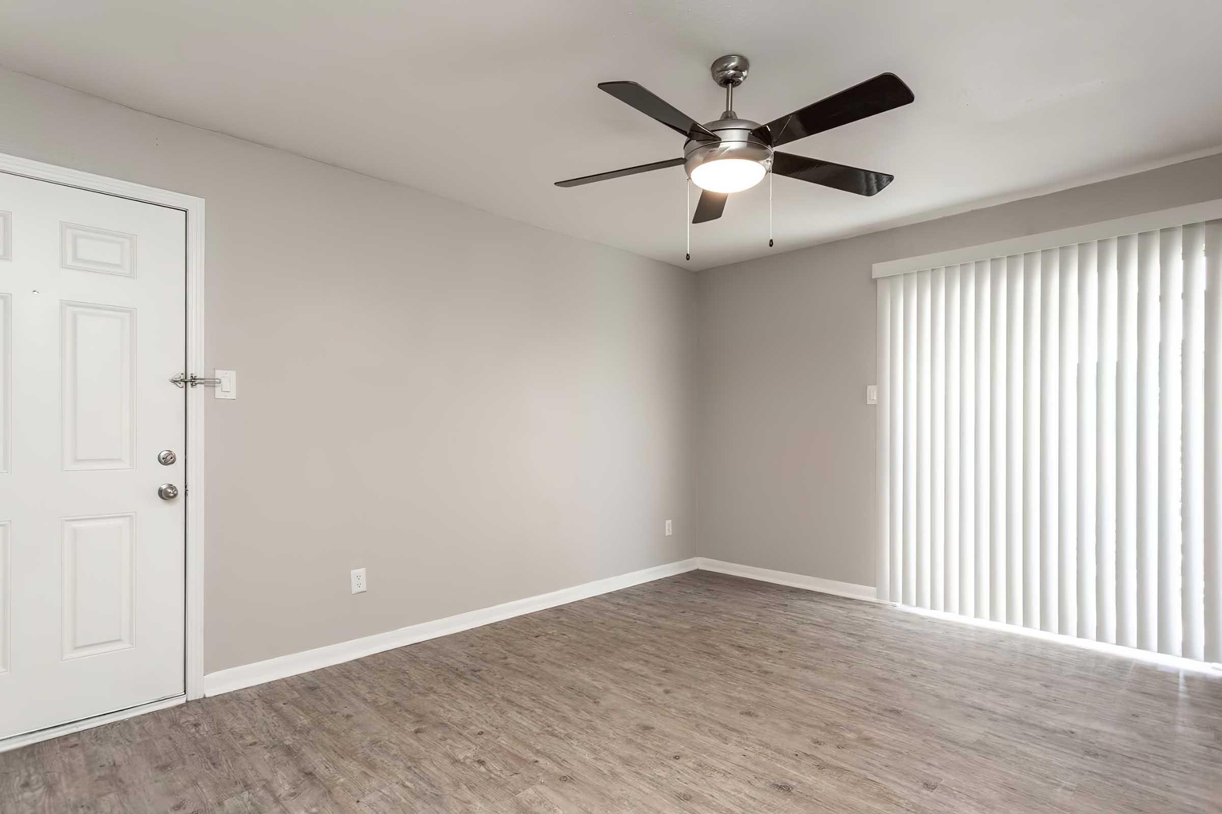 Interior of a room featuring a gray wall, a ceiling fan with a light fixture, a door on the left, and a large window with vertical blinds on the right. The floor has a wood-like finish, and the space appears clean and empty.