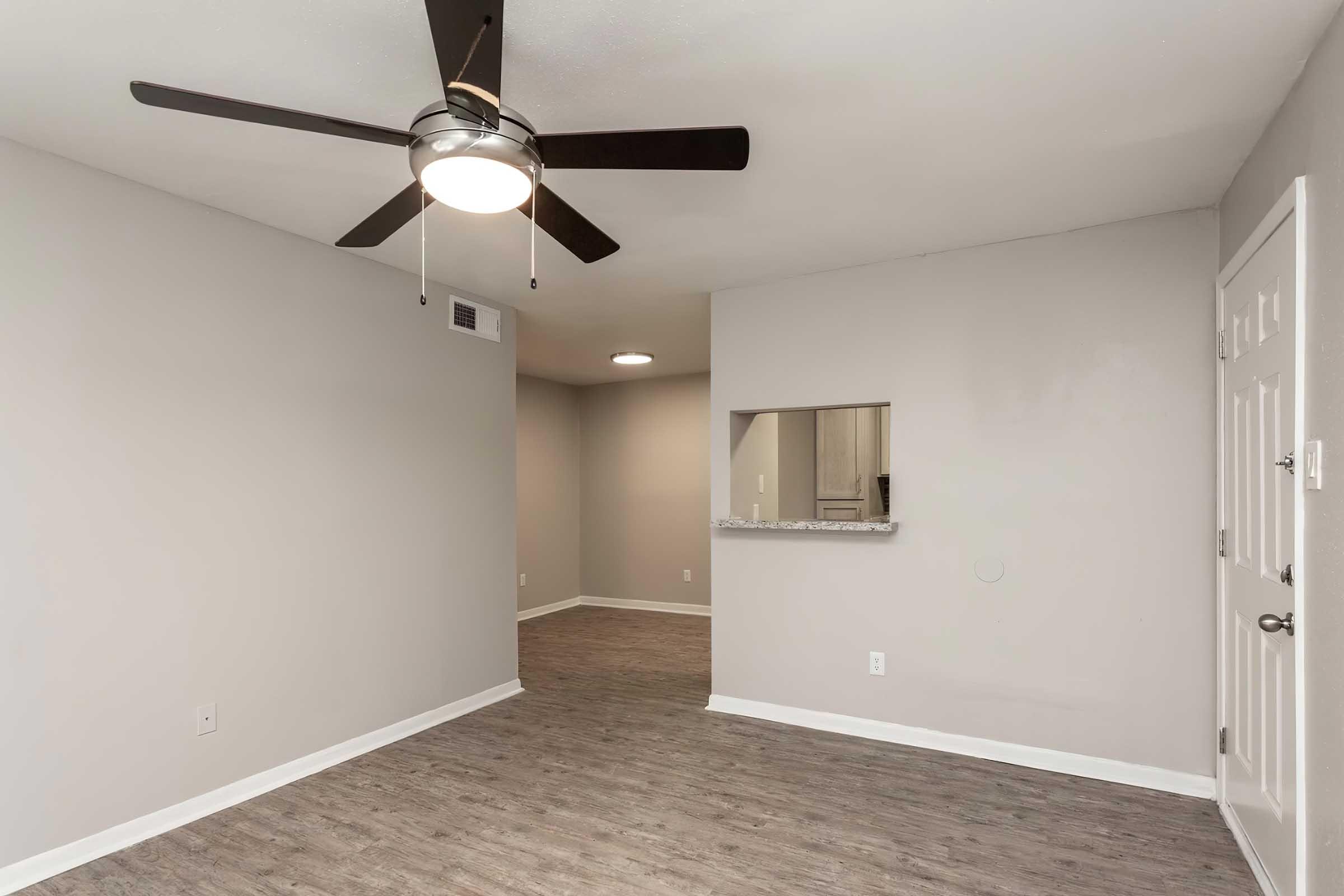 A spacious room featuring a ceiling fan, light gray walls, and a modern design. The floor is covered with a wood-like laminate. A small opening leads to an adjoining area, and there is a door on the right side. The overall atmosphere is bright and open, suitable for various furniture arrangements.