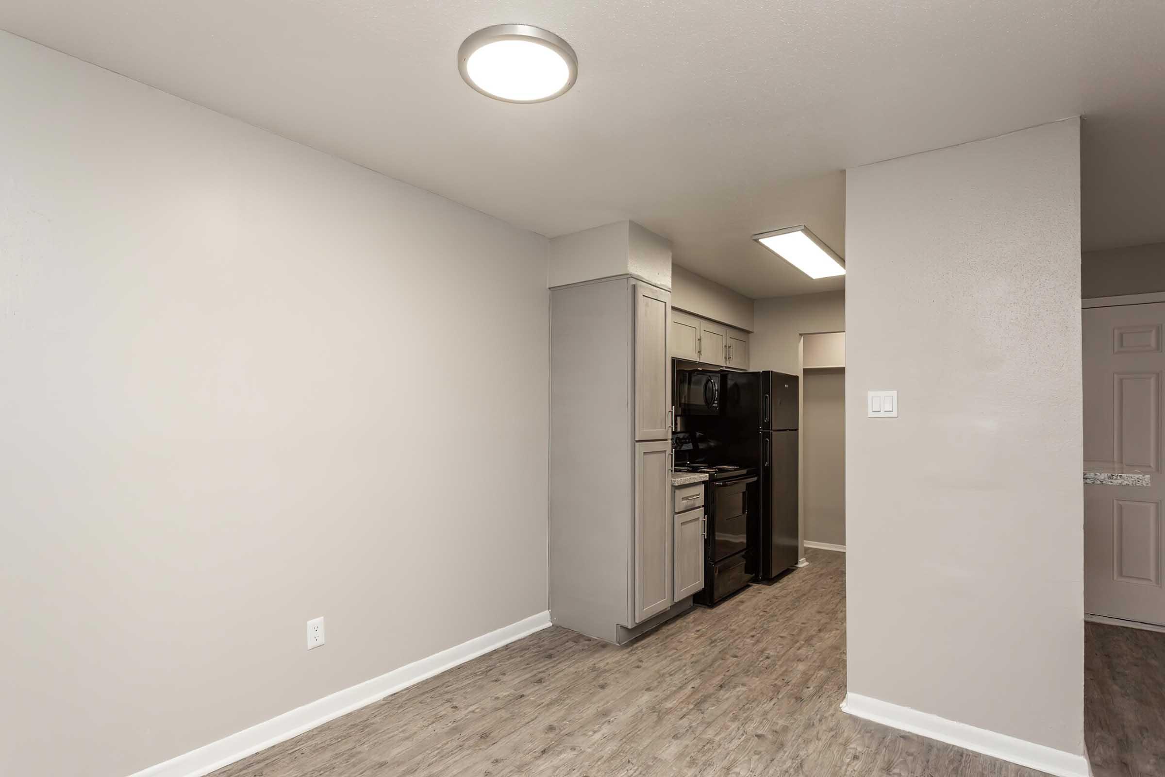Modern apartment interior featuring a light gray color palette. A small kitchen area with black appliances, including a stove and refrigerator, is visible. The flooring appears to be a light wood laminate, and there is a ceiling light fixture providing illumination. The space feels open and clean.
