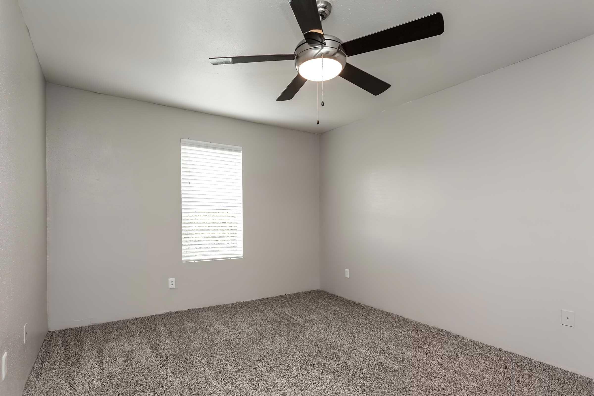 A minimalist room featuring gray walls and carpet. The space includes a window with blinds, allowing natural light in, and a ceiling fan. The room appears empty, providing a blank canvas for furniture and decoration.