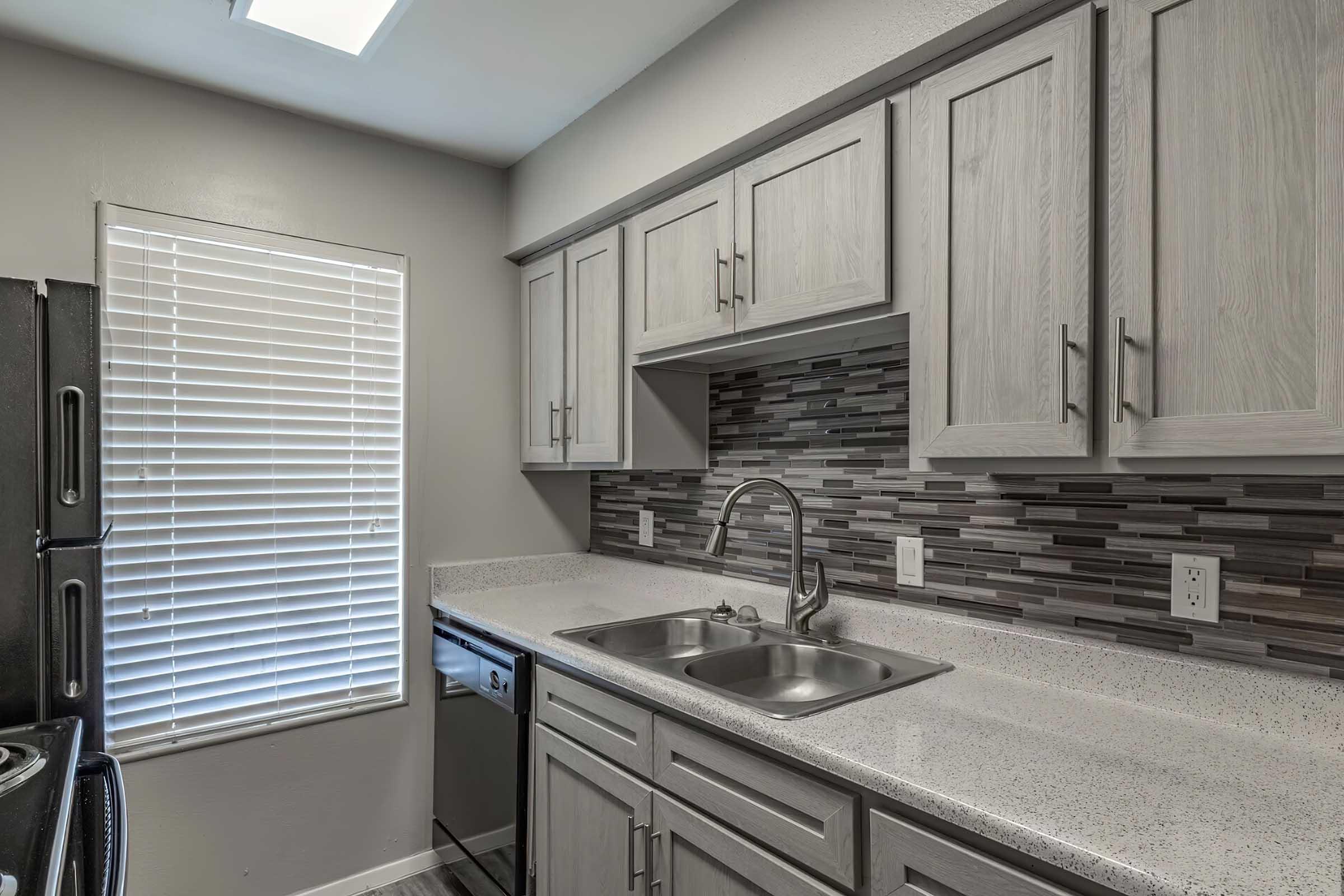 Modern kitchen featuring light gray cabinets, a textured backsplash in shades of gray and black, stainless steel sink with dual faucets, and a large window with white blinds allowing natural light. Countertops are speckled gray granite, and appliances include a black refrigerator and dishwasher.
