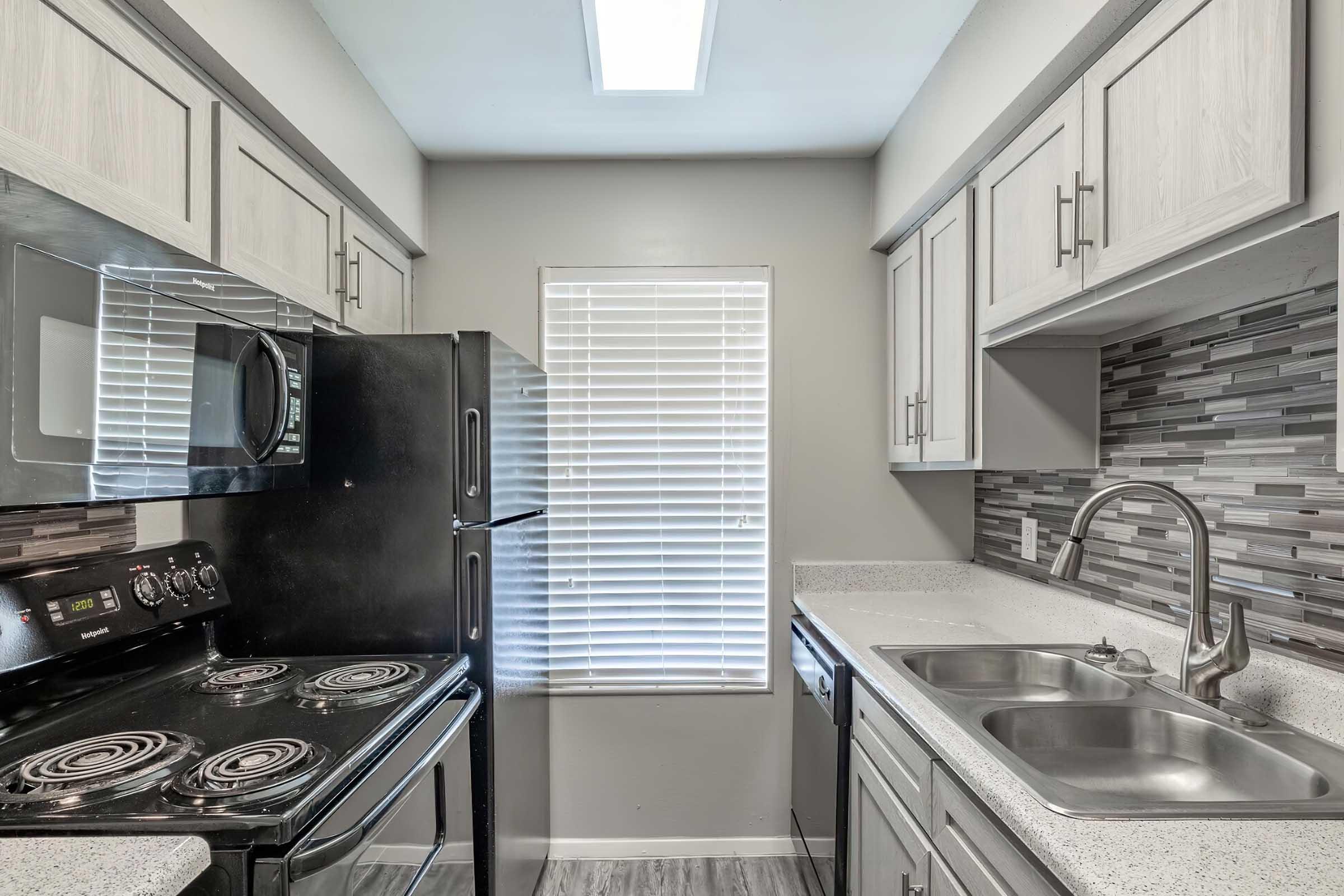 A modern kitchen featuring a black refrigerator, an electric stove, and a double sink. The cabinetry is light-colored, and there is a tile backsplash with gray and black tones. A window with blinds allows natural light into the space. The countertops are gray and have a smooth finish.