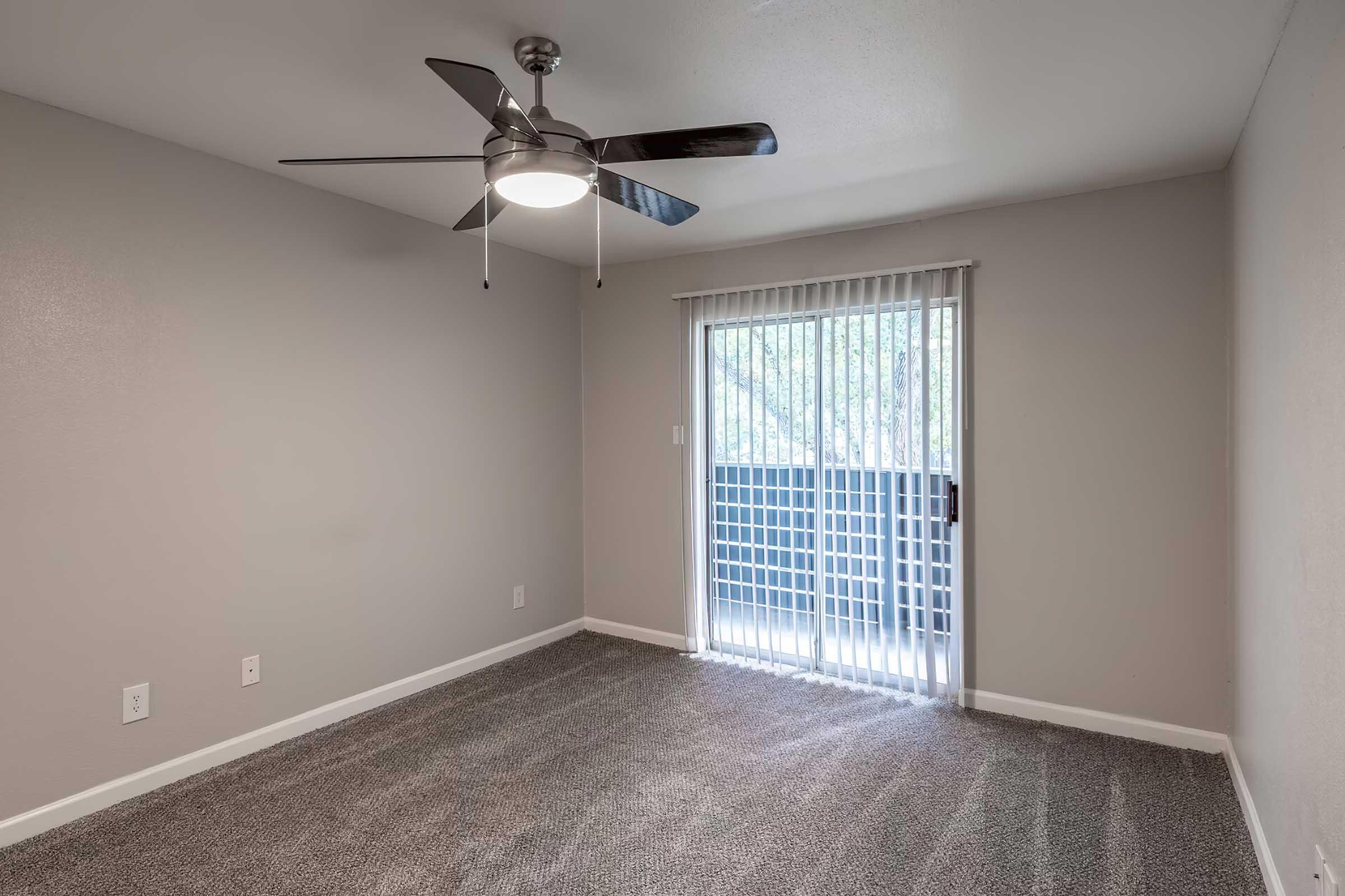 Empty room featuring gray walls and carpeted flooring, with a ceiling fan overhead. A sliding glass door with vertical blinds leads to a balcony. Natural light is softly illuminating the space, creating a bright and open atmosphere.
