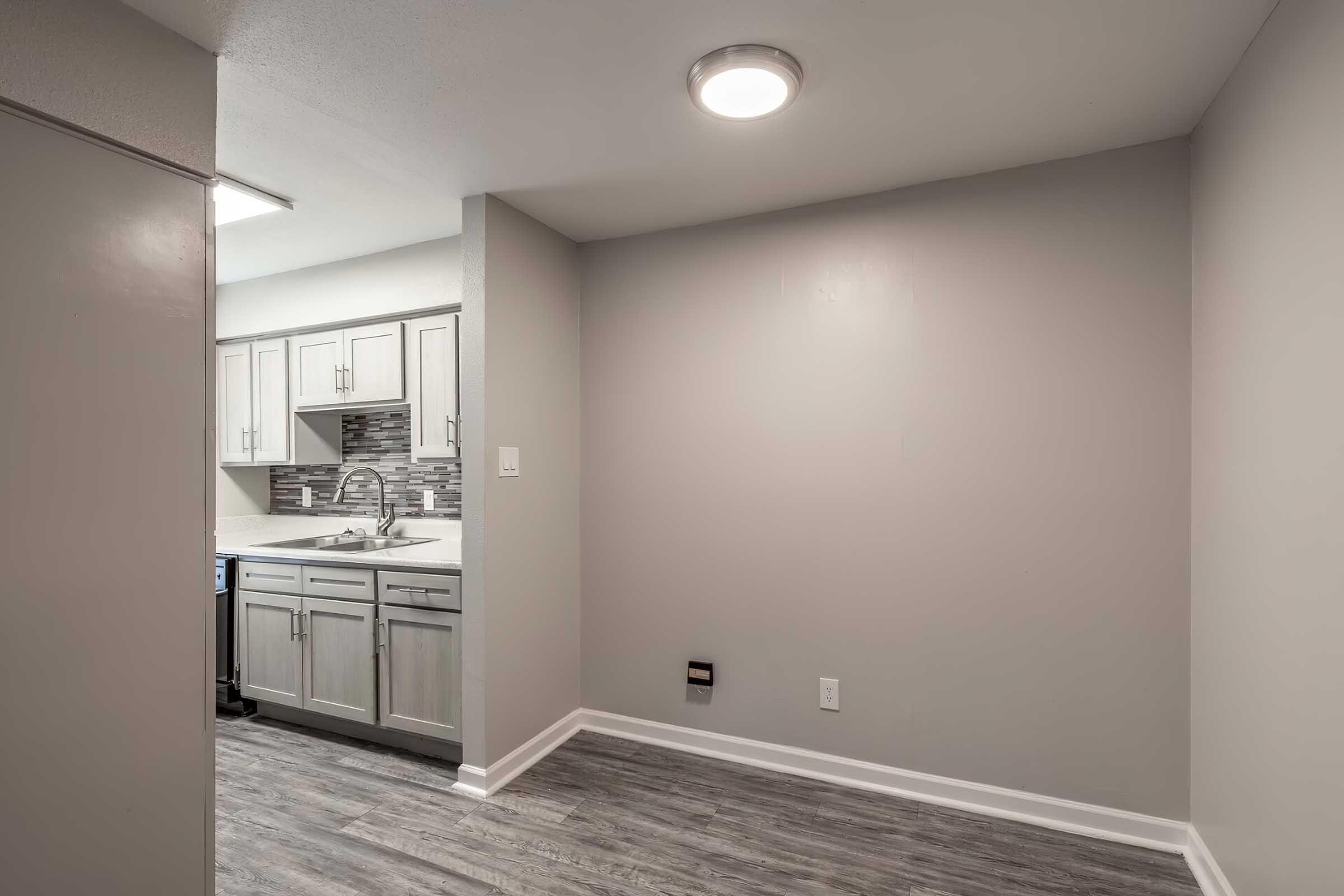 A view of a modern kitchen from a small, empty room. The kitchen area features light gray cabinets with a tile backsplash, a stainless steel sink, and a countertop. The floor is a wood-like laminate, and the walls are painted light gray, with a ceiling light illuminating the space.