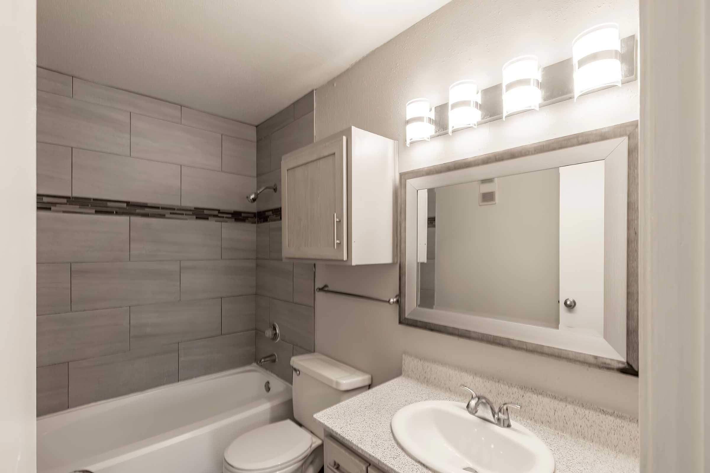 Modern bathroom featuring a bathtub with a shower, a toilet, and a sink with a granite countertop. The walls are tiled in neutral gray tones, and there is a large mirror above the sink. Bright lighting is provided by four wall-mounted light fixtures. Storage is available in a gray cabinet.