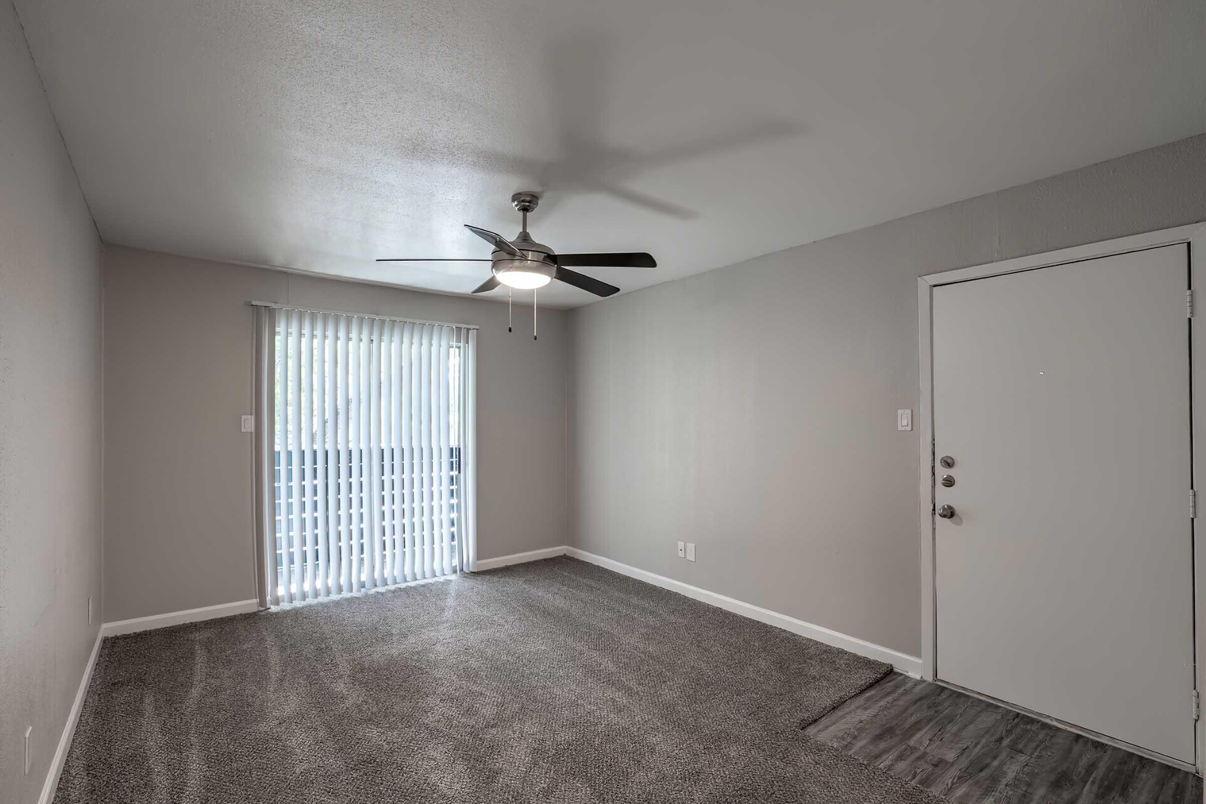 Empty room with light gray walls and carpet, featuring a ceiling fan and vertical blinds on a sliding glass door. A front door is visible on the right side of the image. The space appears spacious and well-lit, suitable for living or office use.