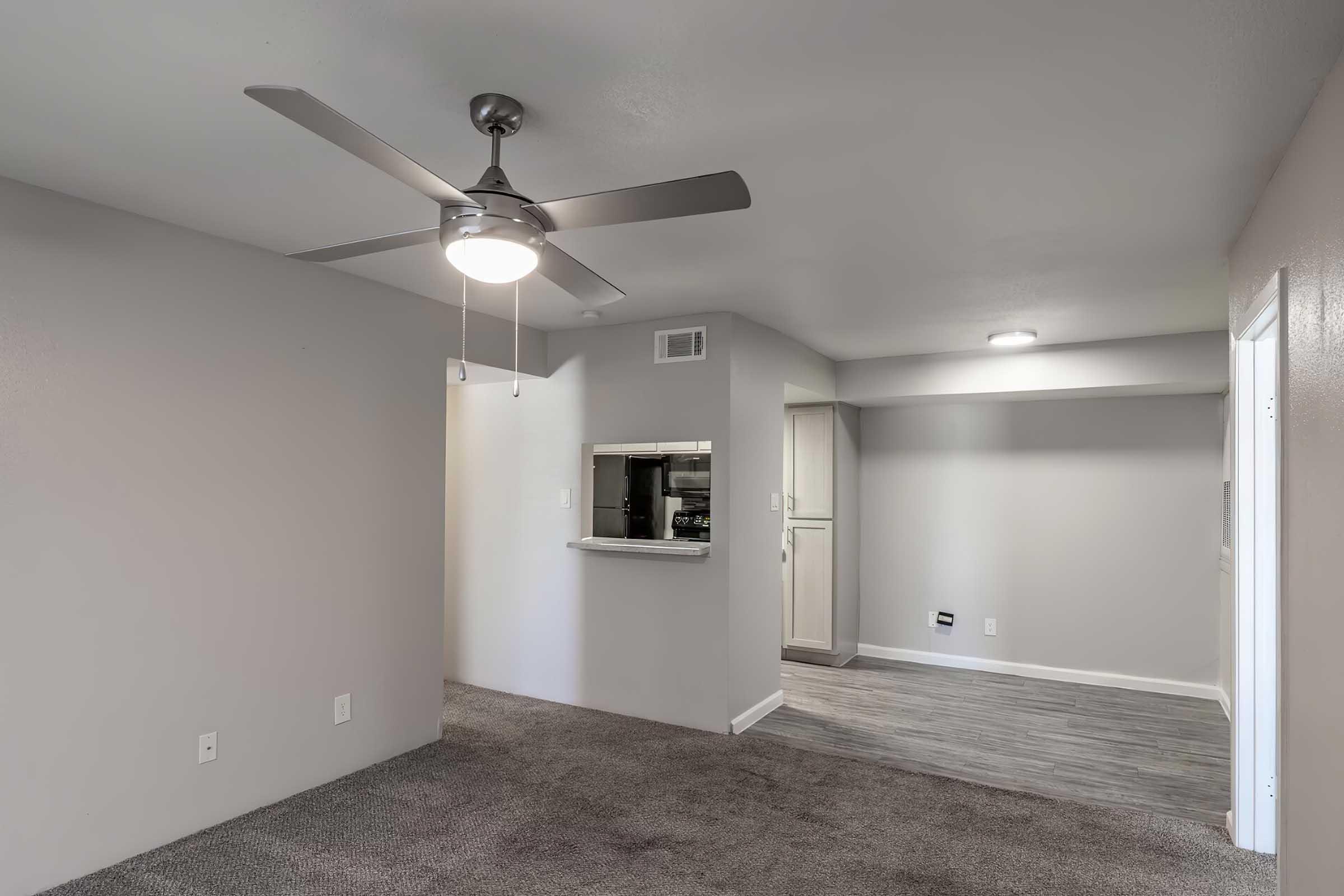 A view of a modern living space featuring light gray walls, a ceiling fan, and carpeted flooring. To the left, there is a small bar area leading to the kitchen. The kitchen has light-colored cabinets, and the right side shows an open area with laminate flooring. The overall atmosphere is bright and inviting.