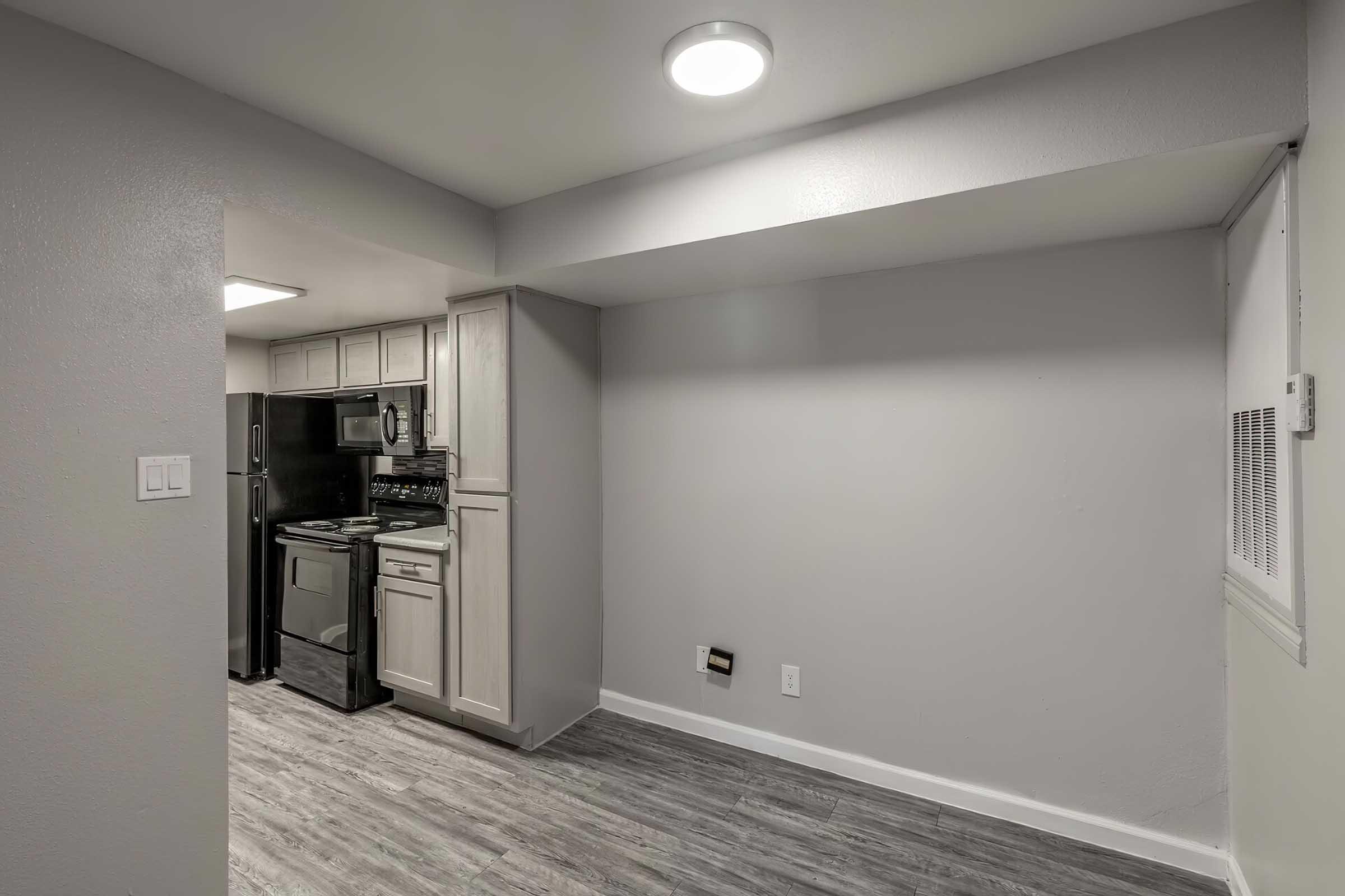 A modern kitchen with gray walls and laminate flooring. It features a black refrigerator and oven, white cabinets, and a ceiling light. A small wall vent is visible, and the space is well-lit with natural light coming from a window.