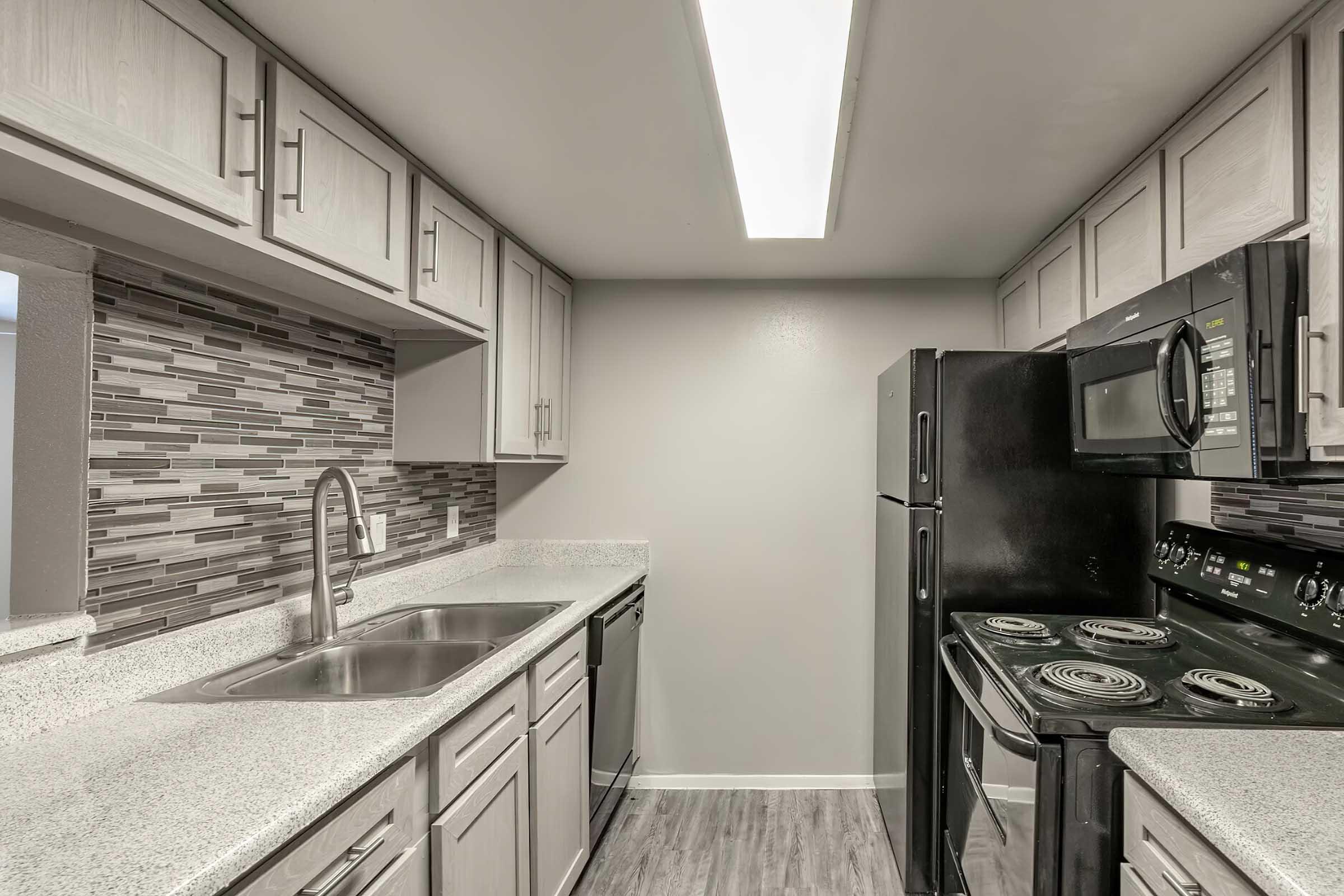 Modern kitchen featuring light gray cabinets, a double sink, black appliances including a microwave and stove, and a decorative tile backsplash. The space has a bright, spacious feel with ample cabinetry and a streamlined design.