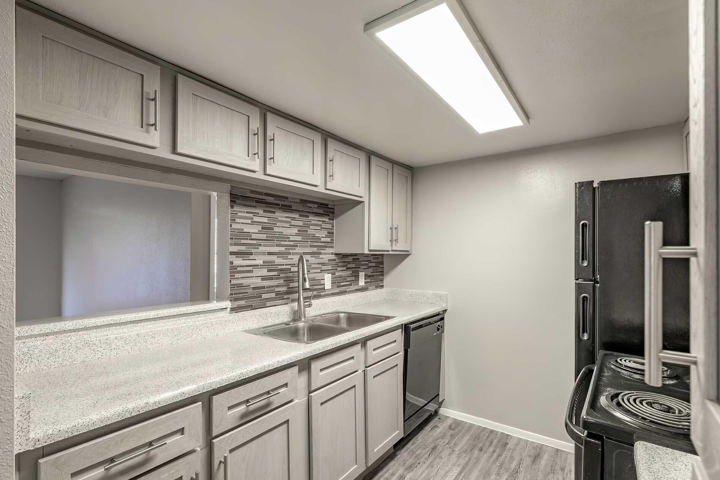 Modern kitchen featuring light gray cabinetry, a stainless steel sink, a black refrigerator, and an electric stove. The backsplash is made of patterned tiles, and there is overhead lighting. The floor is a light wood-like laminate, creating a clean, contemporary look.