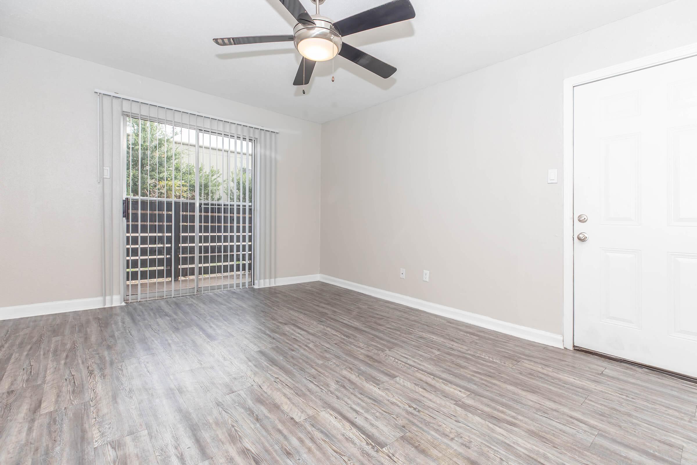 A clean, empty room with modern laminate flooring, featuring a ceiling fan and a sliding glass door that leads to a small outdoor space. The walls are painted in a light neutral color, creating a bright and open atmosphere. A door is visible on the right side of the image.