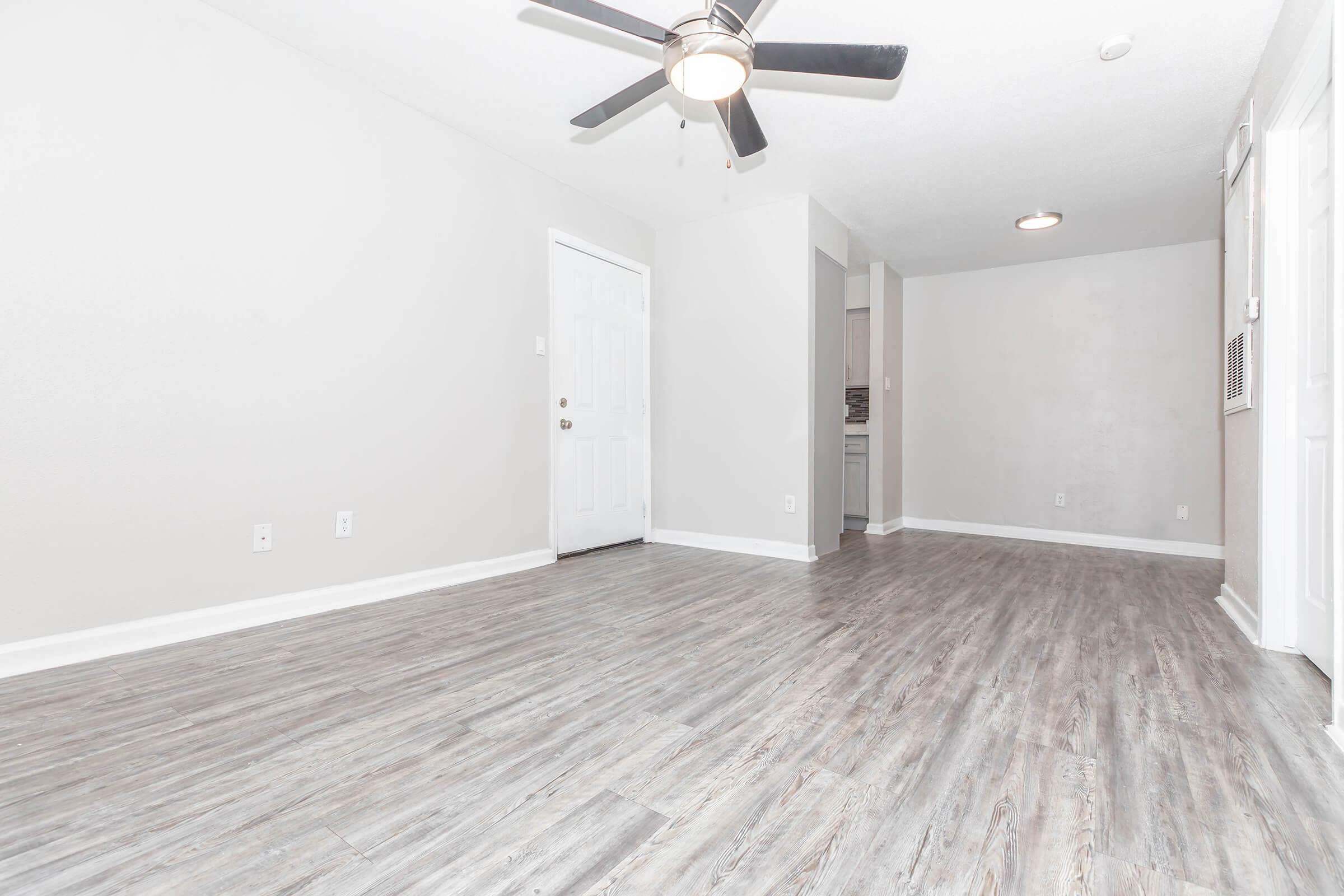 A spacious, empty living room featuring light gray walls and a light wood laminate floor. A ceiling fan is visible above, and there is an open doorway leading to another room. The area is well-lit, with a natural light source illuminating the space.