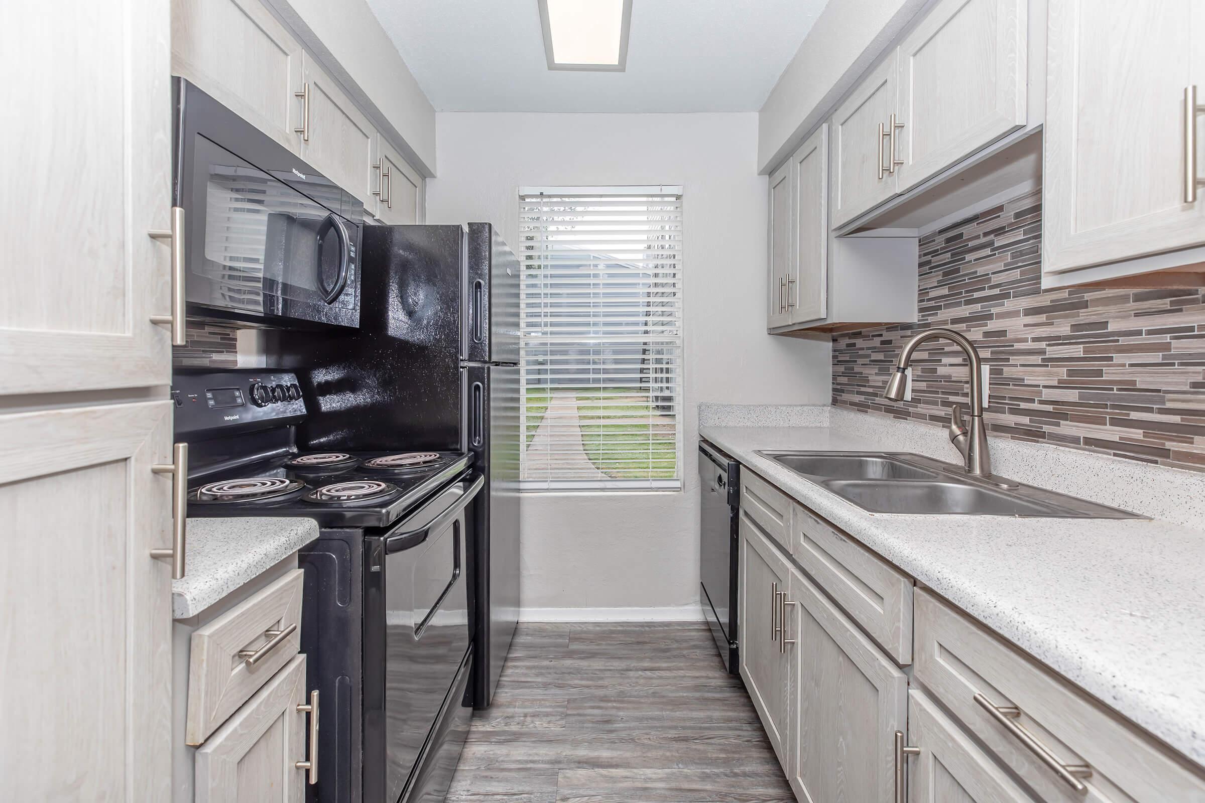 Modern kitchen featuring light wood cabinetry, a black stove and microwave, and a stainless steel sink. The countertop is a smooth gray stone, and there's a decorative tile backsplash. A window with blinds overlooks an outdoor space, and the flooring has a wood-like appearance.