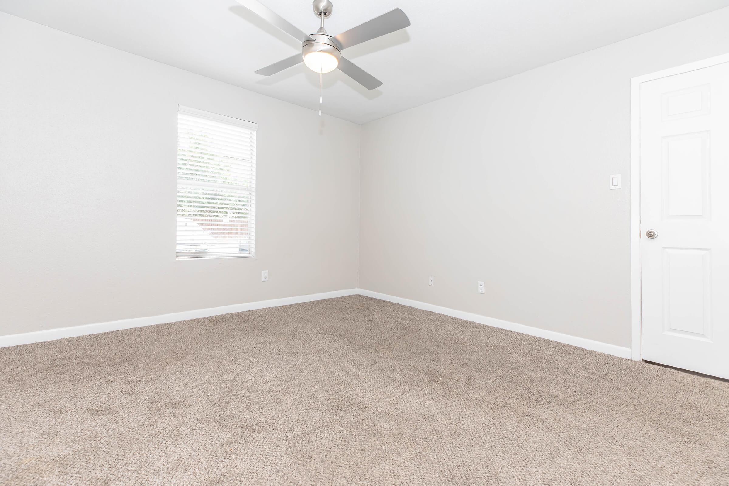 A simple, empty room with light gray walls and beige carpet. It features a ceiling fan, a window with blinds allowing natural light, and a white door on the right side. The space appears clean and well-maintained, making it suitable for furnishing.