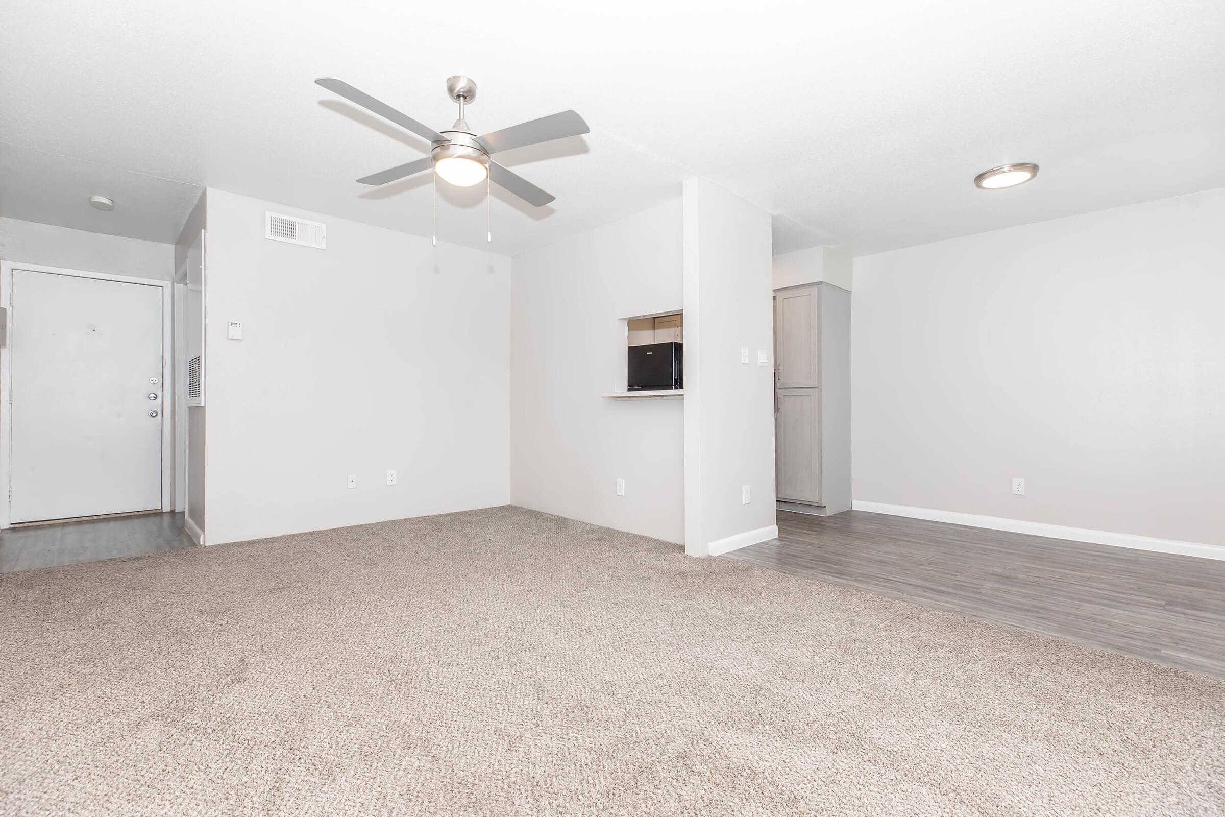 Bright and spacious living area with light-colored walls and a ceiling fan. The room features plush carpet flooring and an open layout leading to a small kitchenette with light cabinetry. A door on the left indicates the entrance, while natural light brightens the space. Ideal for a cozy living environment.