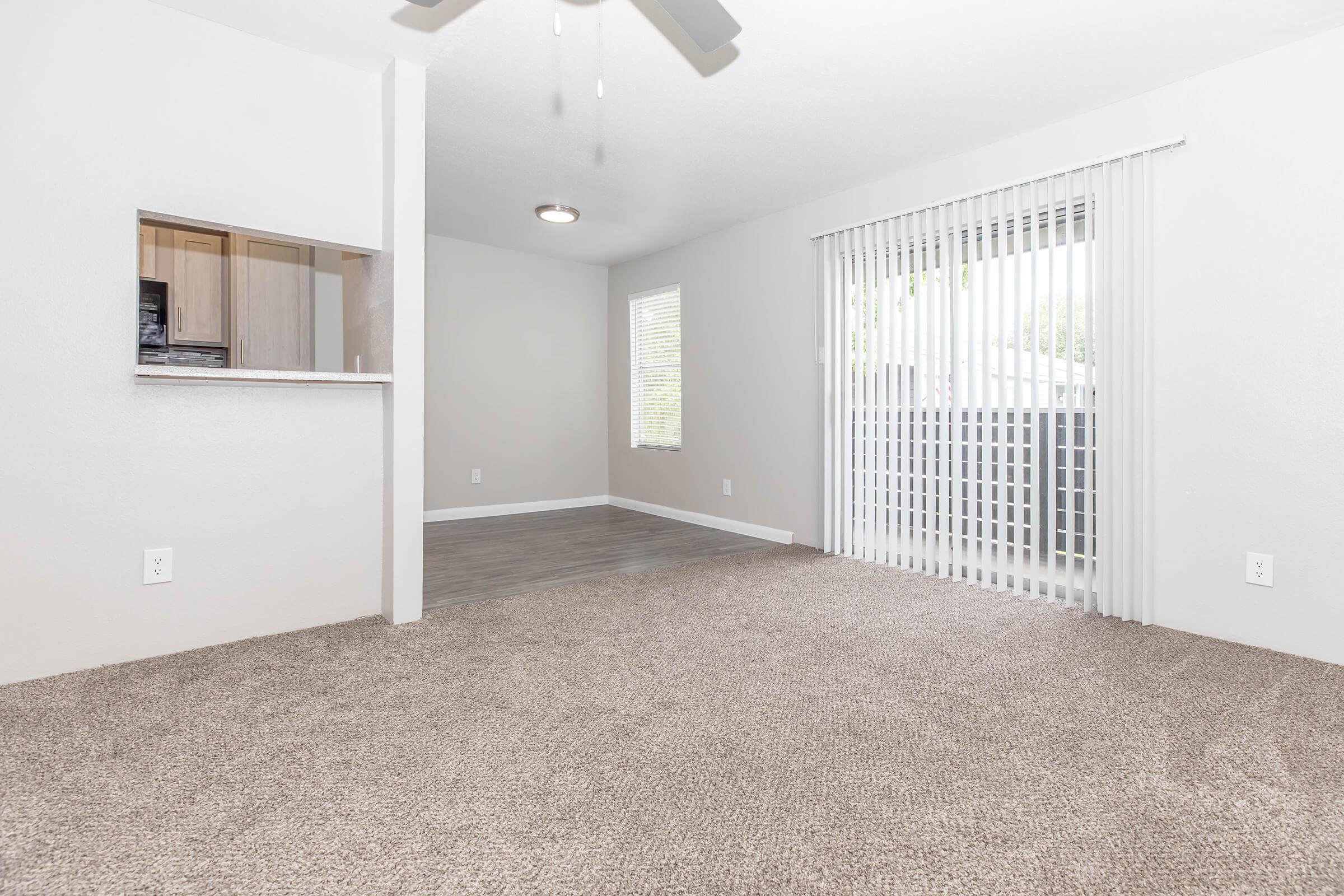 A spacious, well-lit living area featuring light-colored walls and new carpet. A window with vertical blinds allows natural light to enter, and there's a partial view of a kitchen area with a small countertop. The room has a ceiling fan and offers a sense of openness.
