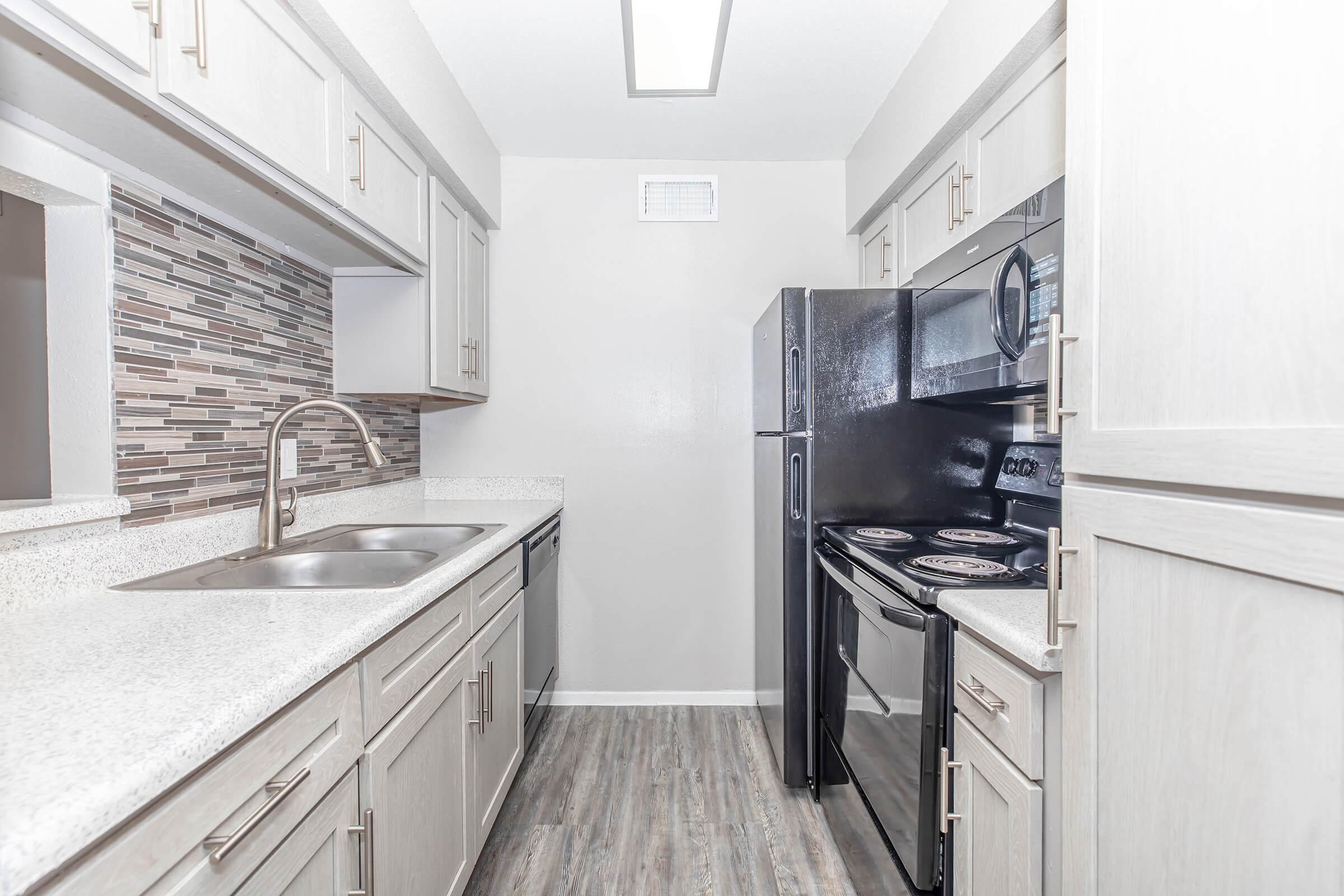 A modern kitchen featuring light-colored cabinets, a double sink, black appliances including a refrigerator and oven, and a stylish backsplash made of mosaic tiles. The floor is a wood-like laminate, and the space has ample natural light coming from a vent above.
