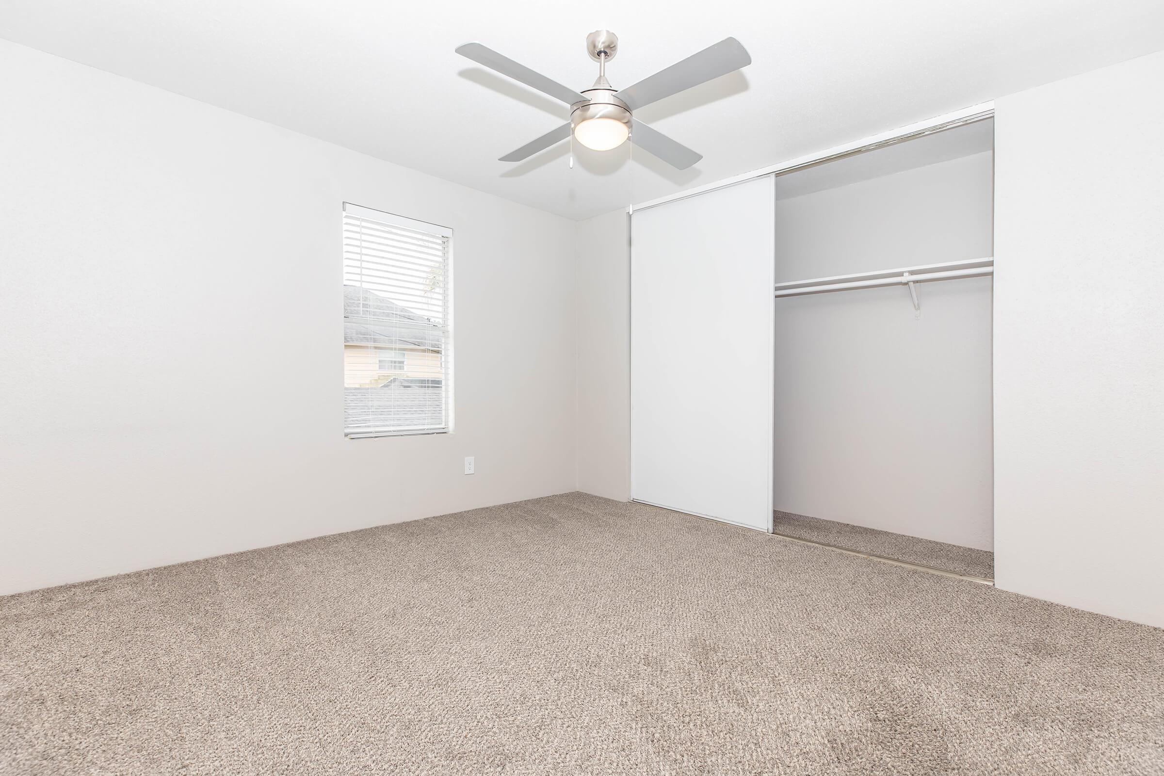 A spacious, empty bedroom featuring light gray walls, a ceiling fan, a single window with blinds, and a large closet with sliding doors. The floor is covered with soft, light-colored carpet, creating a clean and modern look.