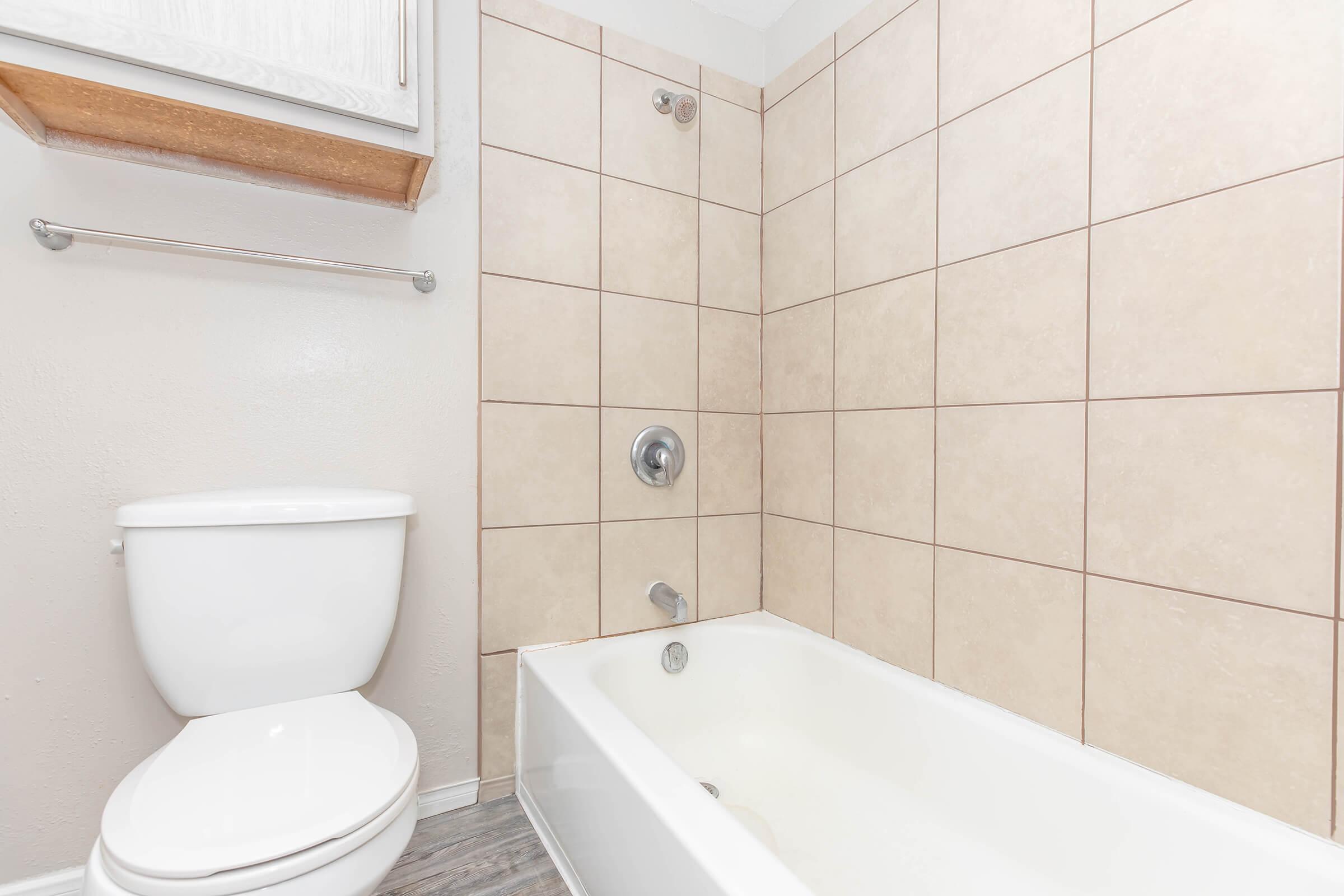 A clean bathroom featuring a white toilet and a bathtub with a showerhead. The walls are tiled with light-colored squares, and there is a wooden shelf above the toilet. The floor has a light wood-like appearance.