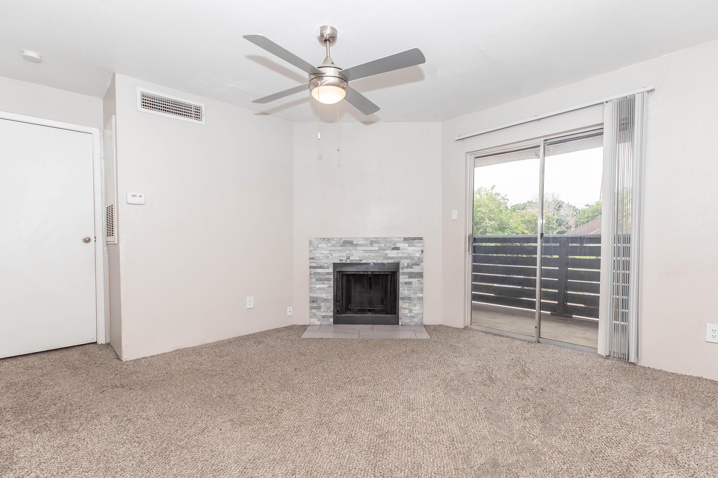 A spacious living room featuring light-colored walls, a ceiling fan, and a carpeted floor. There is a fireplace with a stone surround and a sliding glass door leading to a balcony. Natural light fills the room, creating a warm and inviting atmosphere.