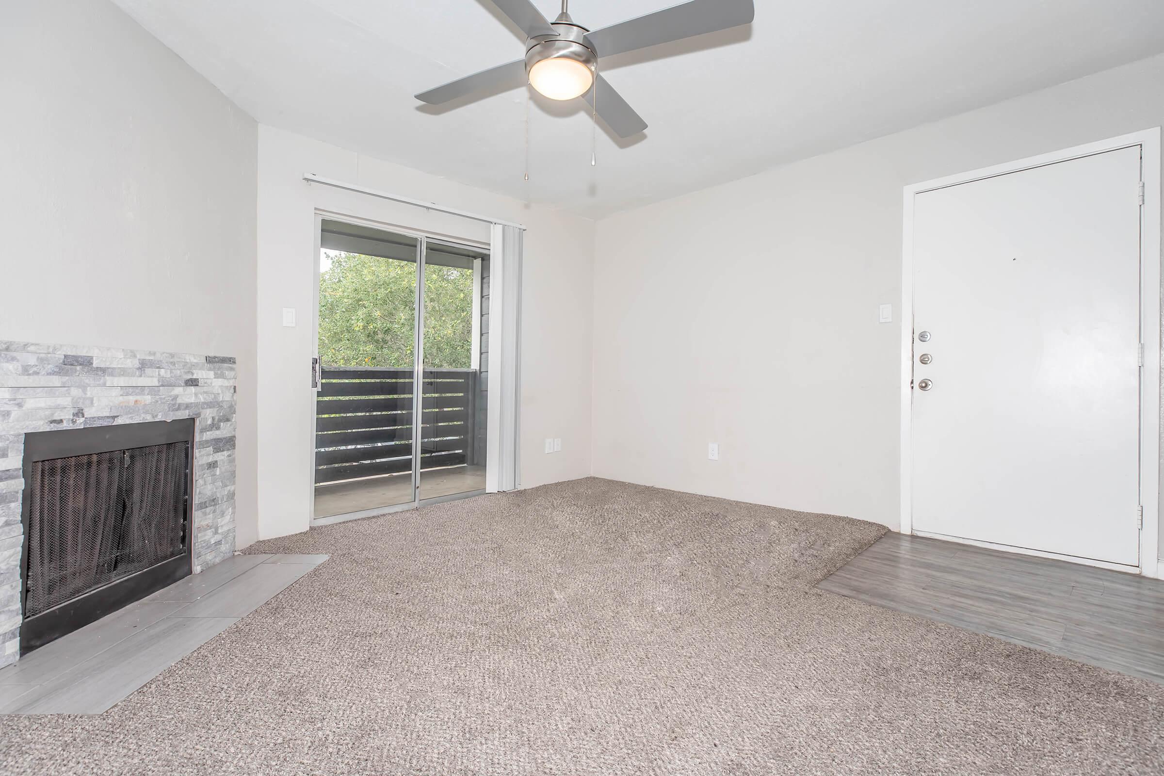 A spacious living room featuring a ceiling fan, carpeted flooring, and a decorative stone fireplace. A sliding glass door leads to a small balcony, while a front door with a security lock is visible. The walls are painted light gray, creating a bright and inviting atmosphere.