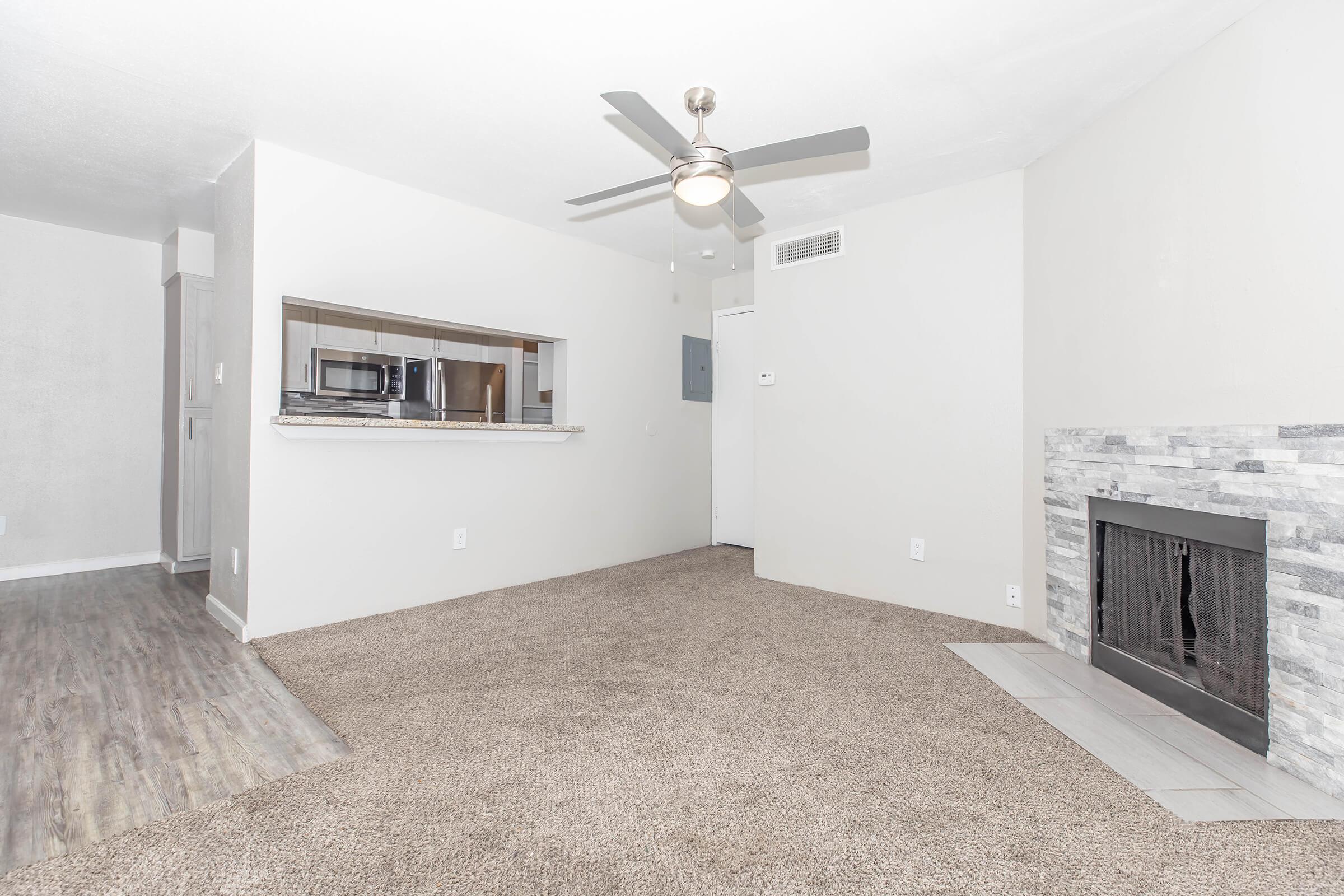 Spacious living room with light-colored walls, plush carpet, and a ceiling fan. A modern fireplace with a gray stone surround is featured, alongside an open view of a kitchen area with a breakfast bar. Natural light enters the room, enhancing the welcoming atmosphere.