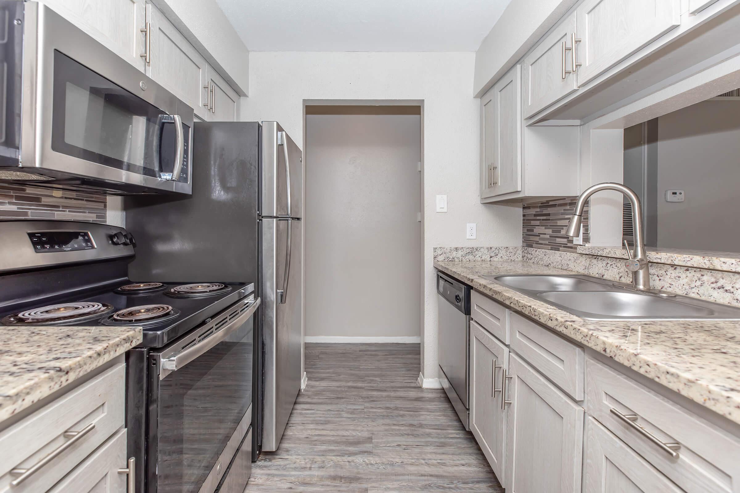 A modern kitchen featuring stainless steel appliances, including a microwave, oven, and refrigerator. The countertops are made of speckled granite, and there is a double sink with a pull-out faucet. Light-colored cabinets provide storage, and the floor is a gray-toned laminate. An open doorway leads to another room.