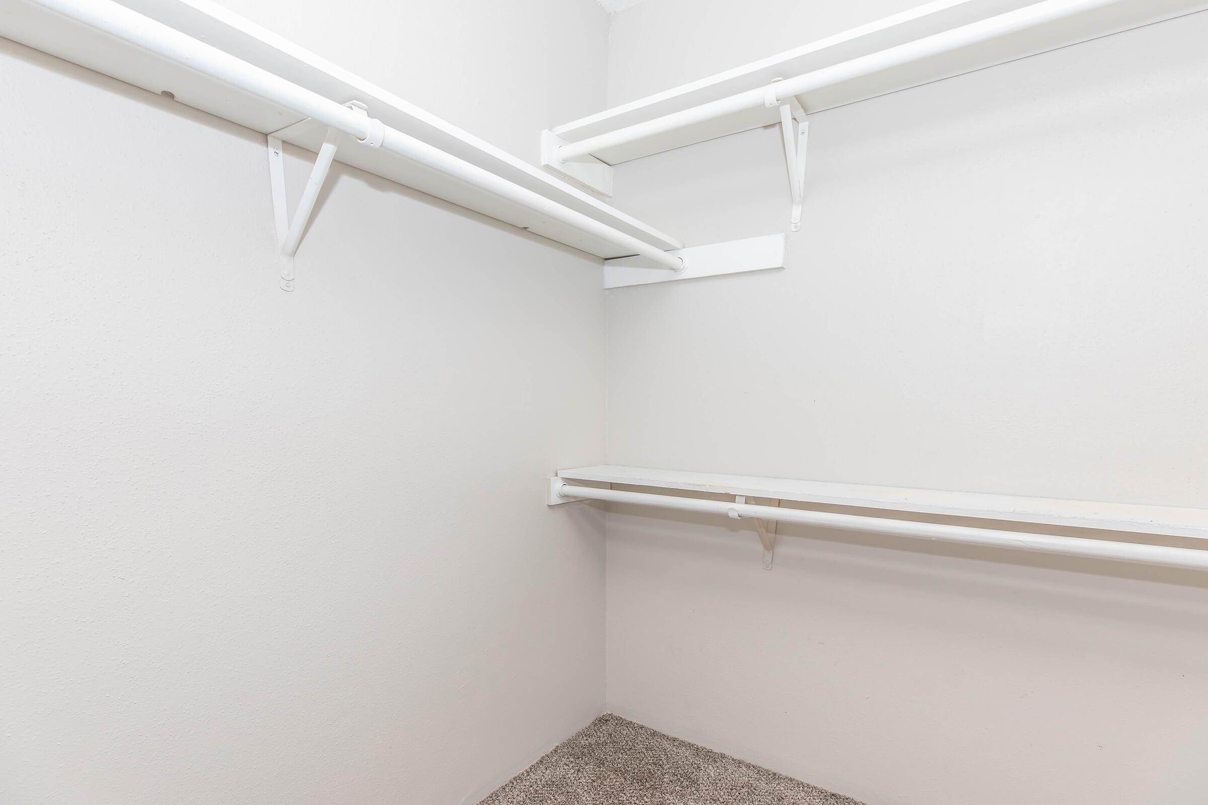 A clean, empty walk-in closet featuring two white shelving units mounted on light-colored walls. The floor is covered with a neutral carpet, creating a spacious and minimalist look.