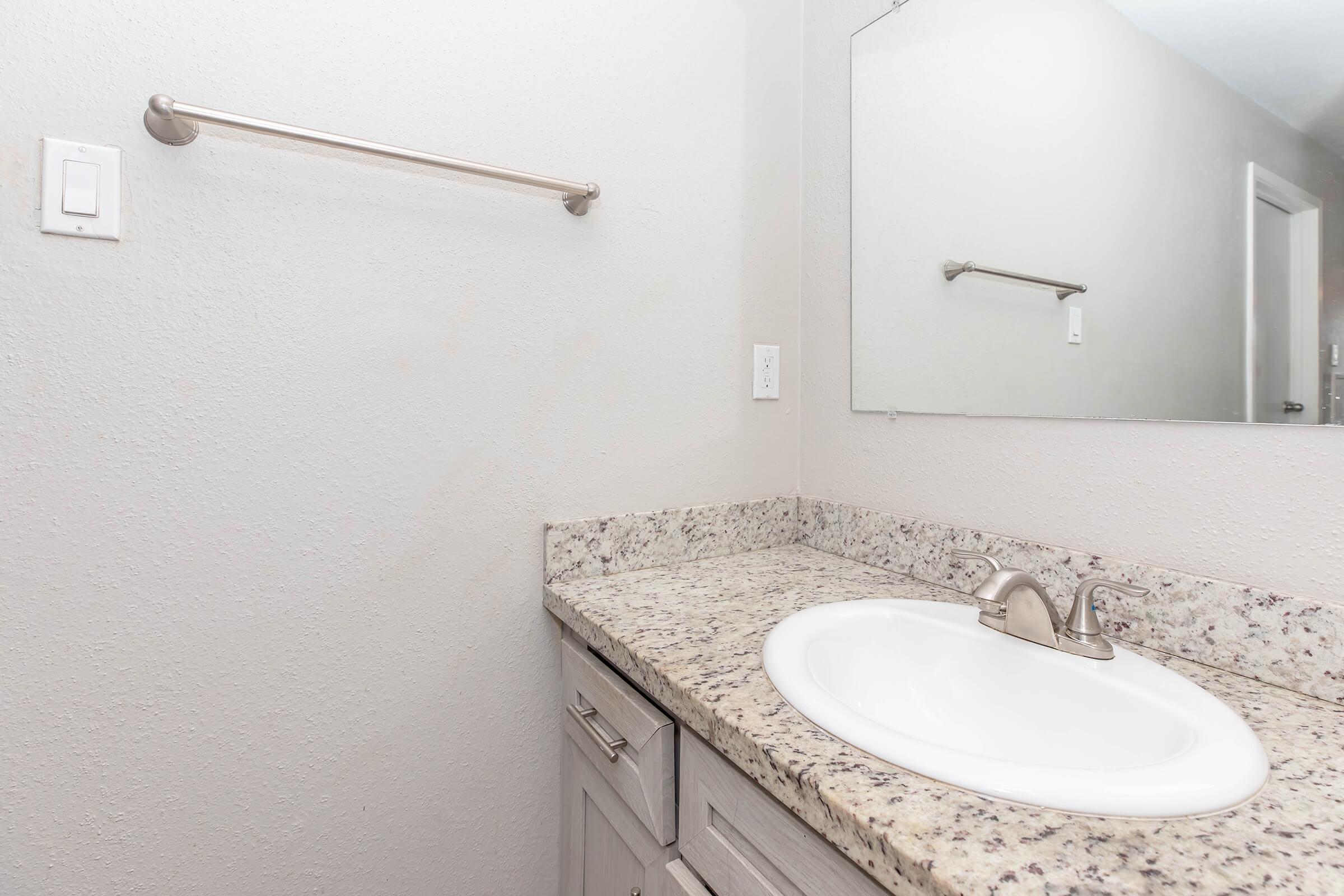 A clean and minimalistic bathroom featuring a light-colored granite countertop with a white sink, a wall-mounted mirror, and a single towel bar. The walls are neutral-toned, creating a bright and spacious appearance. The cabinetry consists of light wood, enhancing the modern look of the space.