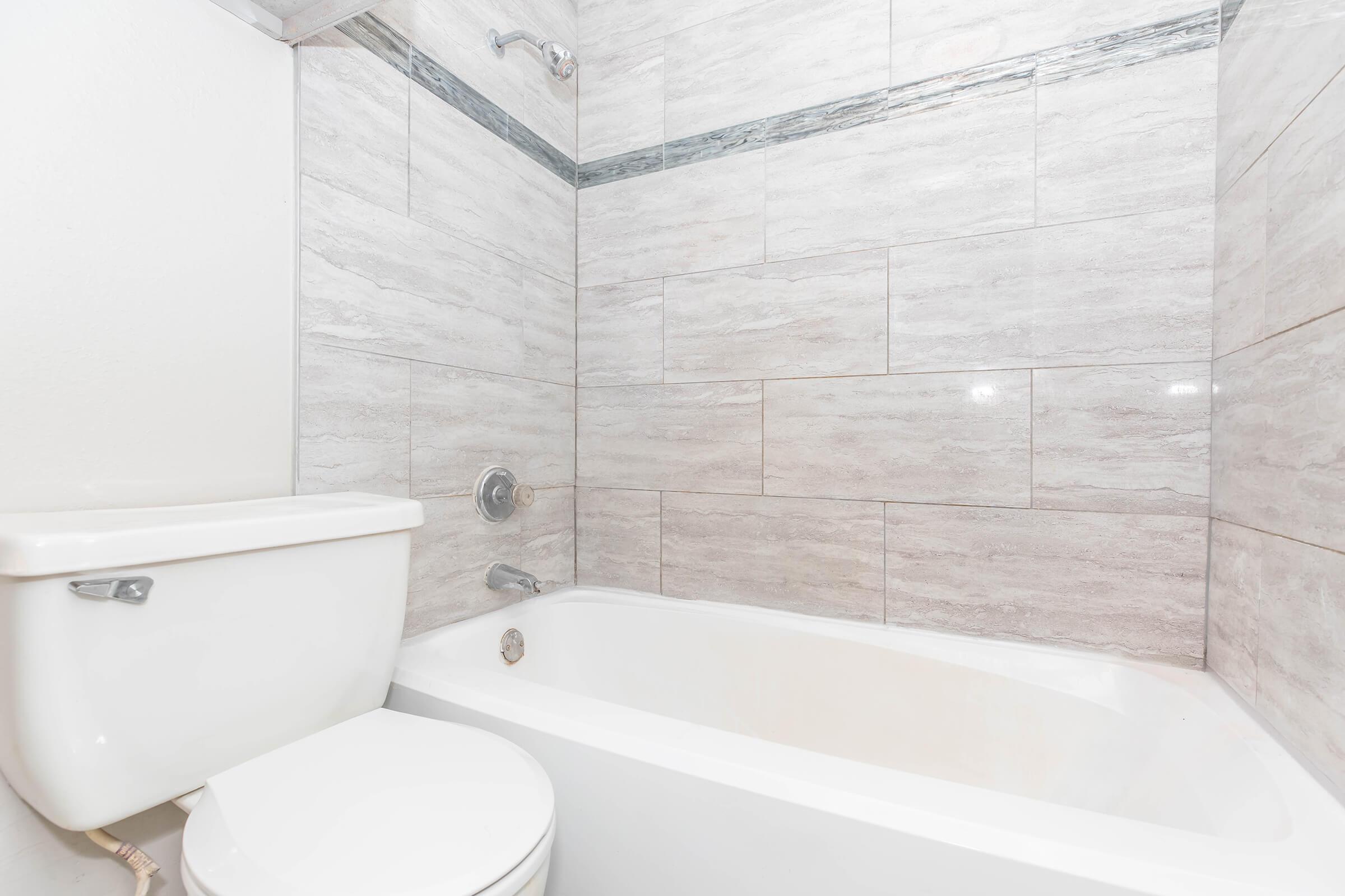 A clean bathroom featuring a white toilet and a bathtub with gray tiled walls. The bathtub is empty, and there's a showerhead above it. The space is well-lit, showcasing modern finishes and a minimalist design.