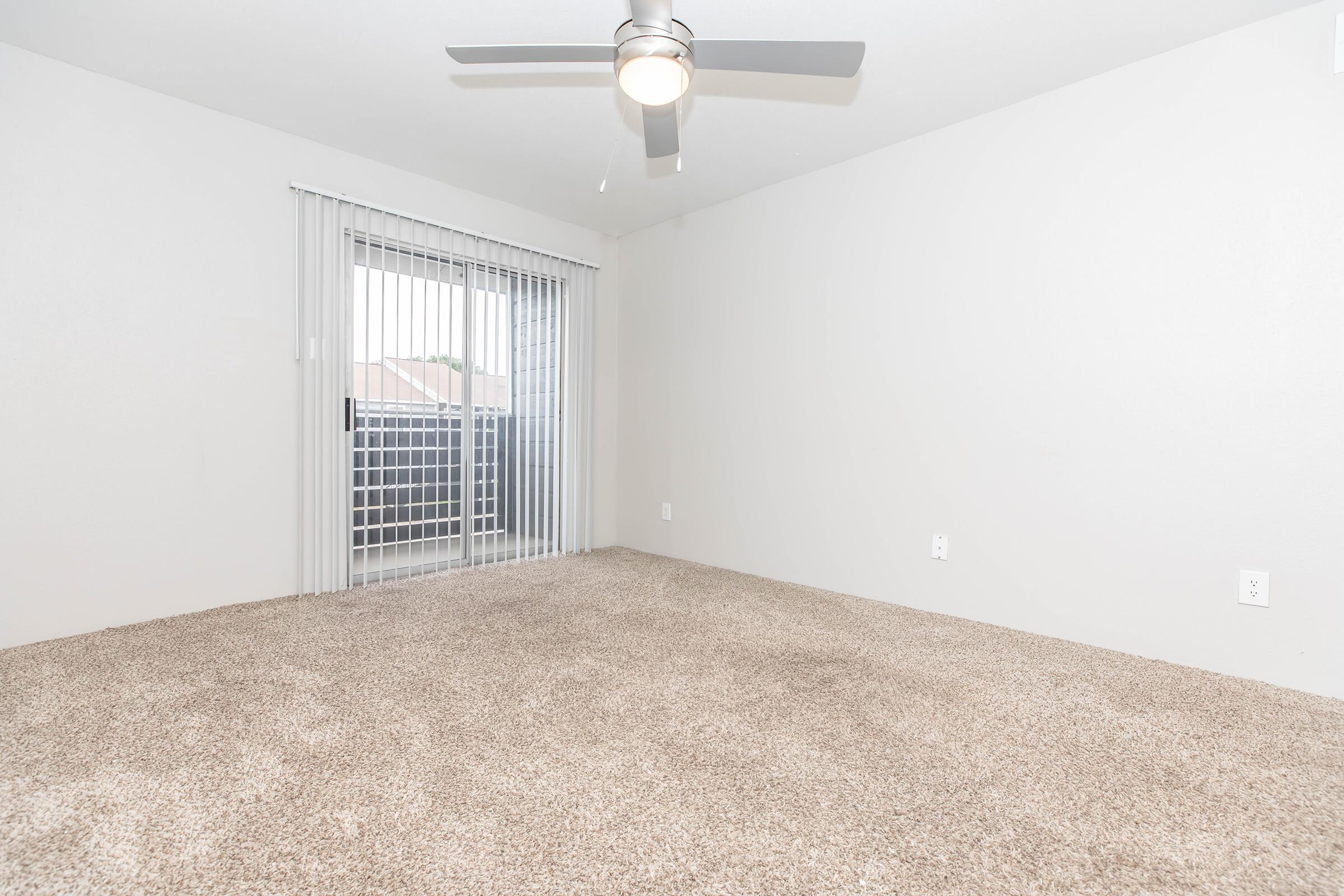 Empty room with light beige carpet, white walls, and a ceiling fan. A sliding glass door with vertical blinds leads to a balcony, allowing natural light into the space. The room is minimally decorated, creating an open and spacious atmosphere.