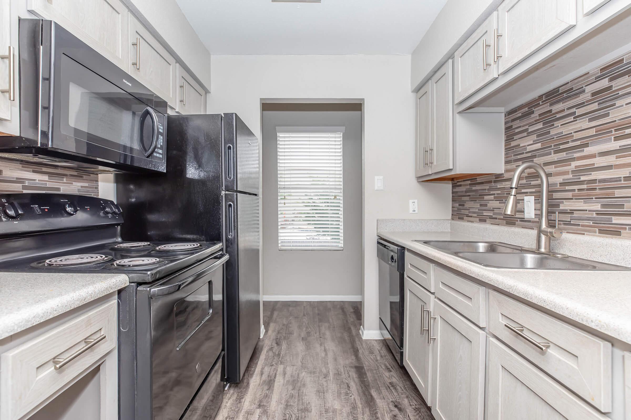 A modern kitchen featuring light gray cabinets, a black refrigerator, and a black stove with a sleek design. The countertop is light-colored, and there is a stainless steel sink. The backsplash consists of multicolored tiles, and a window provides natural light, enhancing the overall brightness of the space.