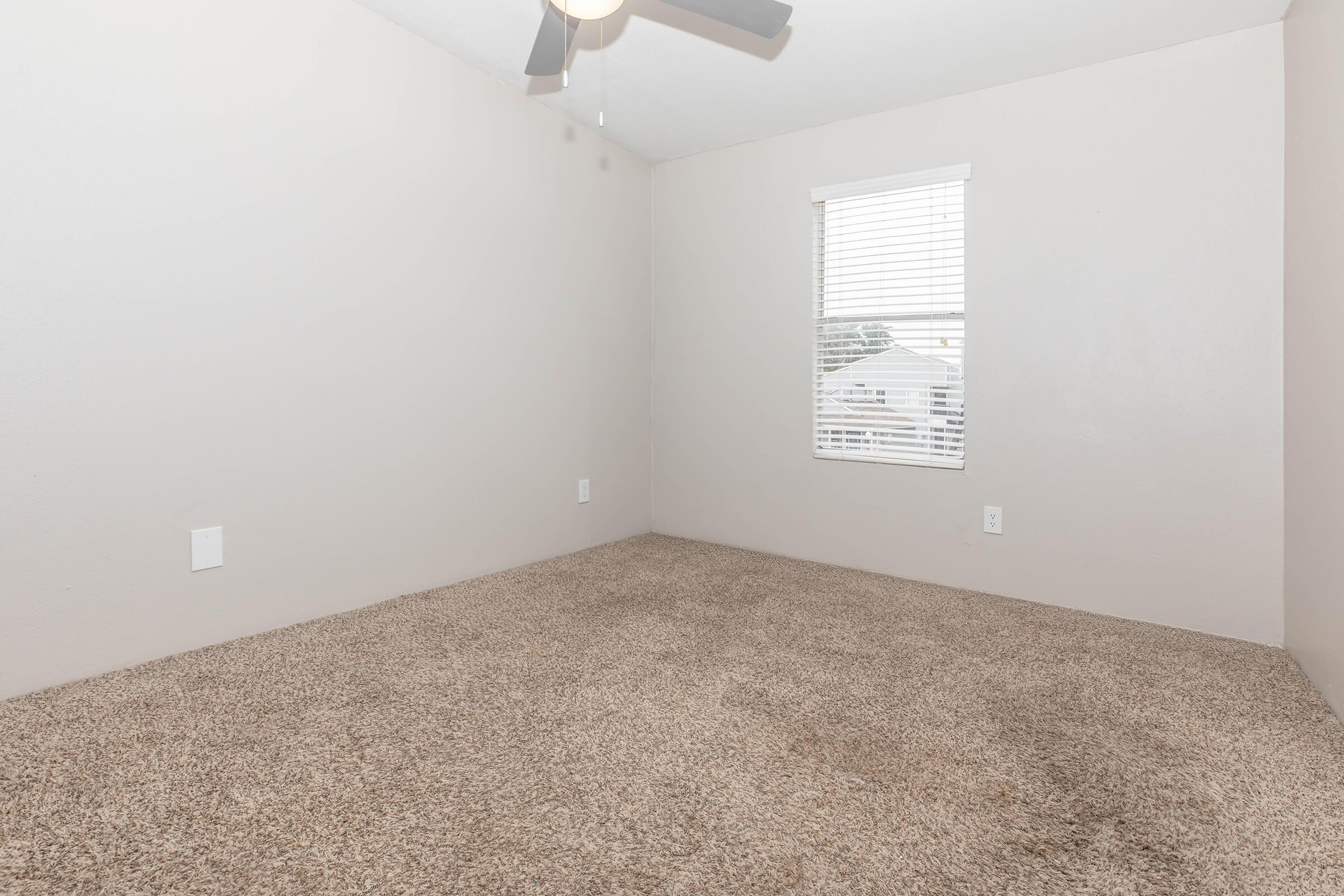 A small, empty room featuring light beige walls, a ceiling fan, and a window with white blinds. The floor is covered in plush beige carpet, contributing to a neutral and spacious feel.