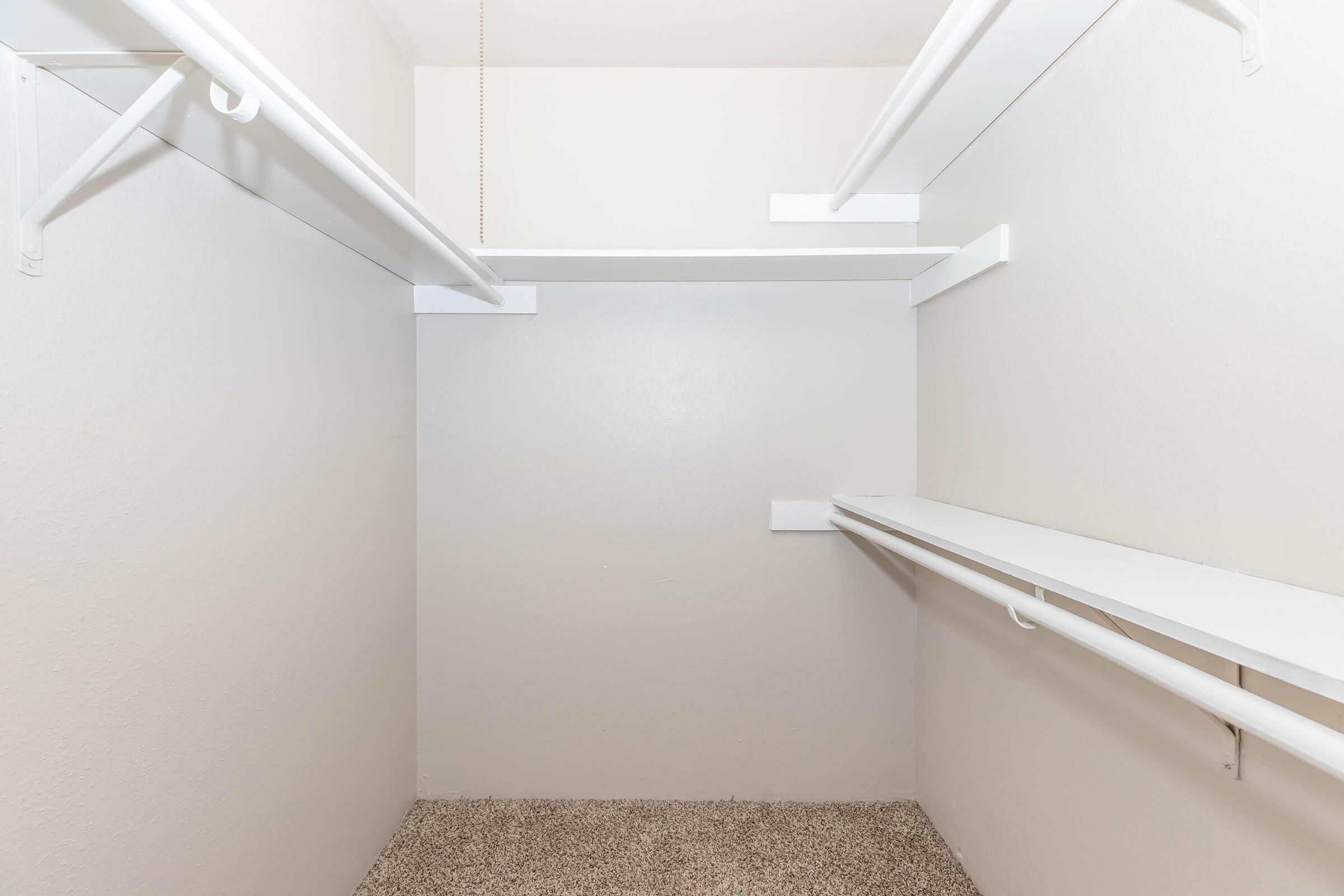 Empty closet with white walls and shelving. The floor is covered in light beige carpet, and there are white horizontal shelves and a hanging rod on either side, creating a spacious and organized appearance. The lighting is bright, enhancing the clean look of the space.