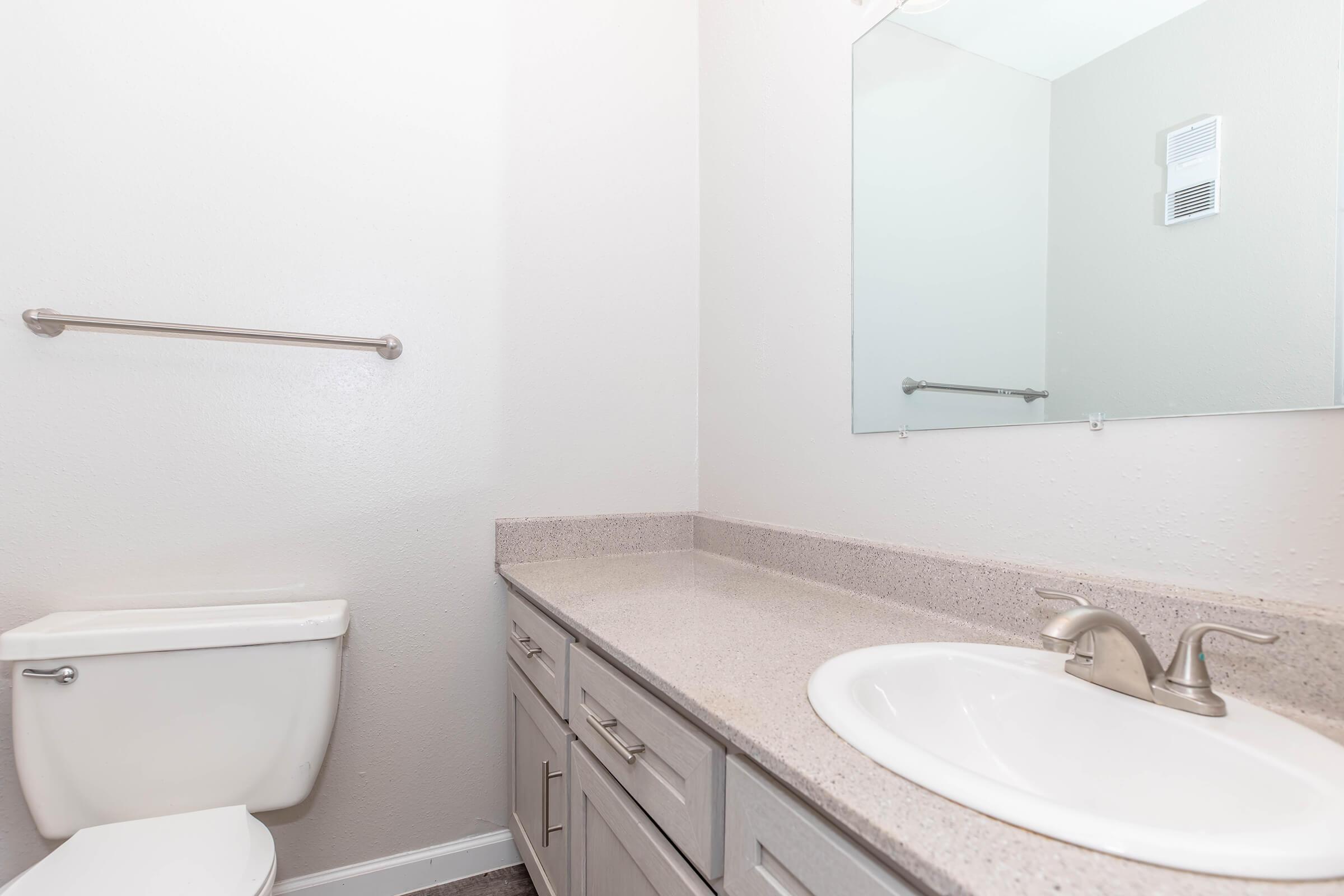 A clean, modern bathroom featuring a white toilet, a light gray countertop with sink, and a large mirror above the sink. The walls are painted in a light color, and there is a towel bar mounted on the wall. The space is well-lit and has a minimalist design.