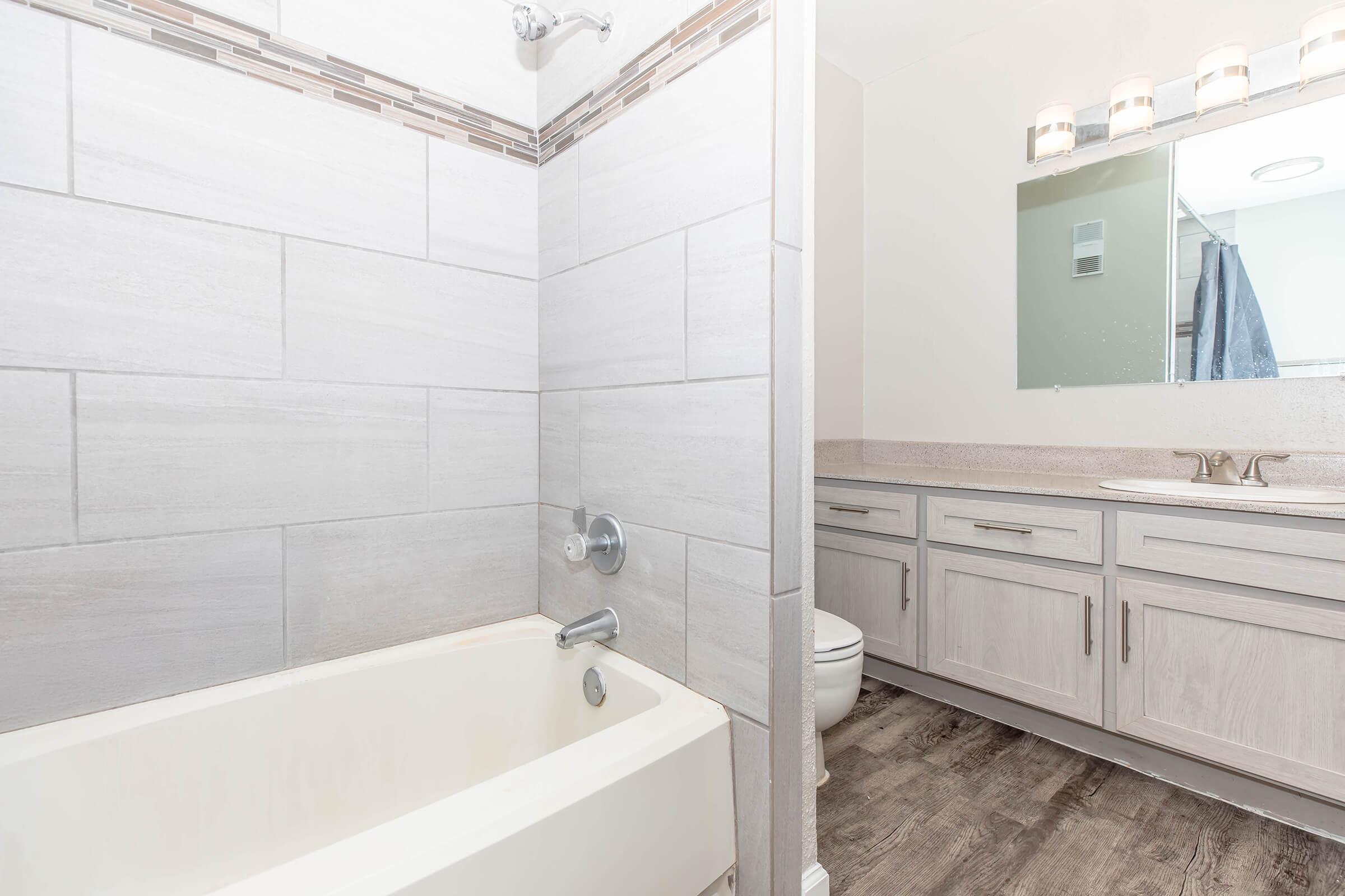 A modern bathroom featuring a shower and bathtub combo with light gray tiled walls. The adjacent area has a double vanity with light wood cabinetry and a large mirror. Neutral-colored flooring and minimal decor contribute to a clean, contemporary aesthetic.