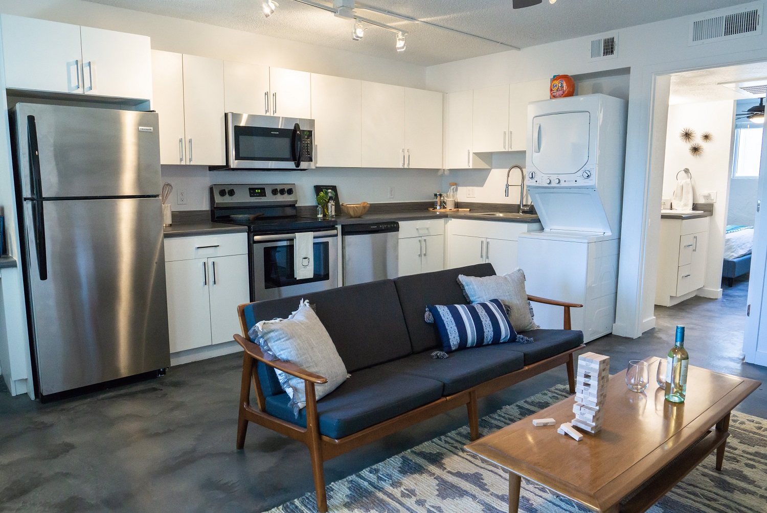 A modern kitchen and living area featuring stainless steel appliances, white cabinetry, a dark countertop, and a comfortable brown sofa with decorative pillows. A wooden coffee table is in front, holding a bottle of wine and a game. A laundry area is visible in the background.