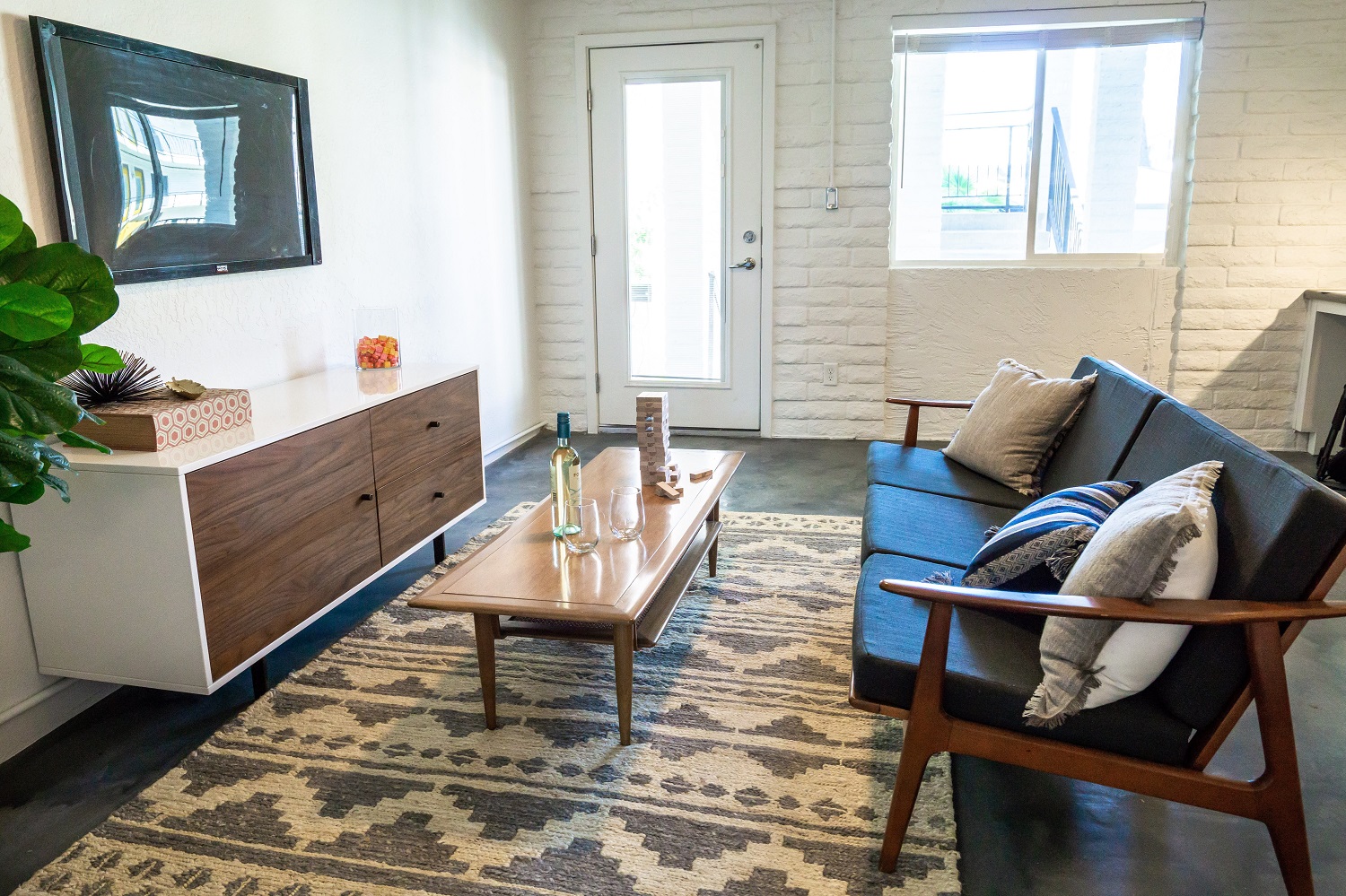 A stylish living room featuring a modern sofa with decorative pillows, a wooden coffee table with a bottle and glasses, and a sideboard. A large window allows natural light to brighten the space. An area rug adds warmth, and a potted plant provides a touch of greenery.