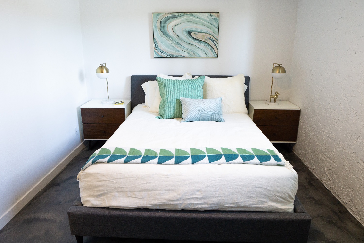 A modern bedroom featuring a neatly made bed with white linens and a decorative teal throw blanket. There are two bedside tables with lamps on either side of the bed, and a wall art piece above the bed with a swirling design in teal and beige tones. The room has light-colored walls and a simple, contemporary aesthetic.