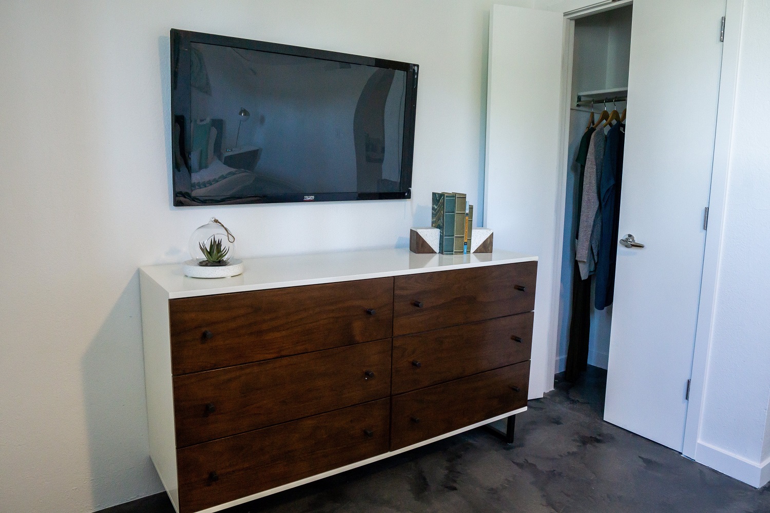 A modern bedroom interior featuring a wooden dresser with six drawers, a small potted plant on top, and a wall-mounted television above. To the right, an open closet reveals a couple of hanging garments, and a door leads to another space. The walls are painted white, creating a spacious and bright atmosphere.