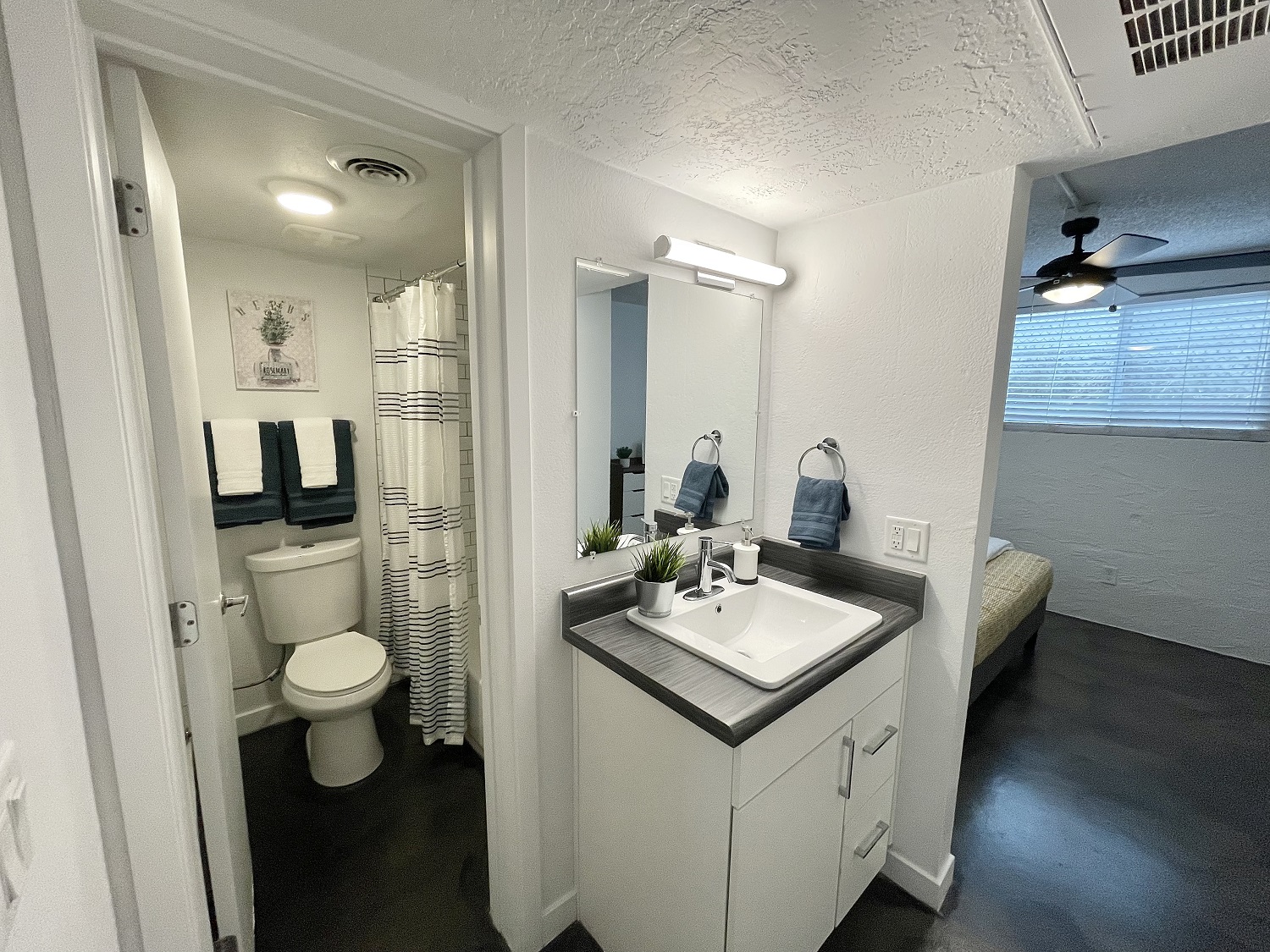 A modern bathroom featuring a white vanity with a sink, mirror, and toiletries. A shower curtain with stripes is visible, along with towels hanging neatly. In the background, an adjacent room shows a bed with a neatly made quilt and a ceiling fan. The space is well-lit with neutral-colored walls.