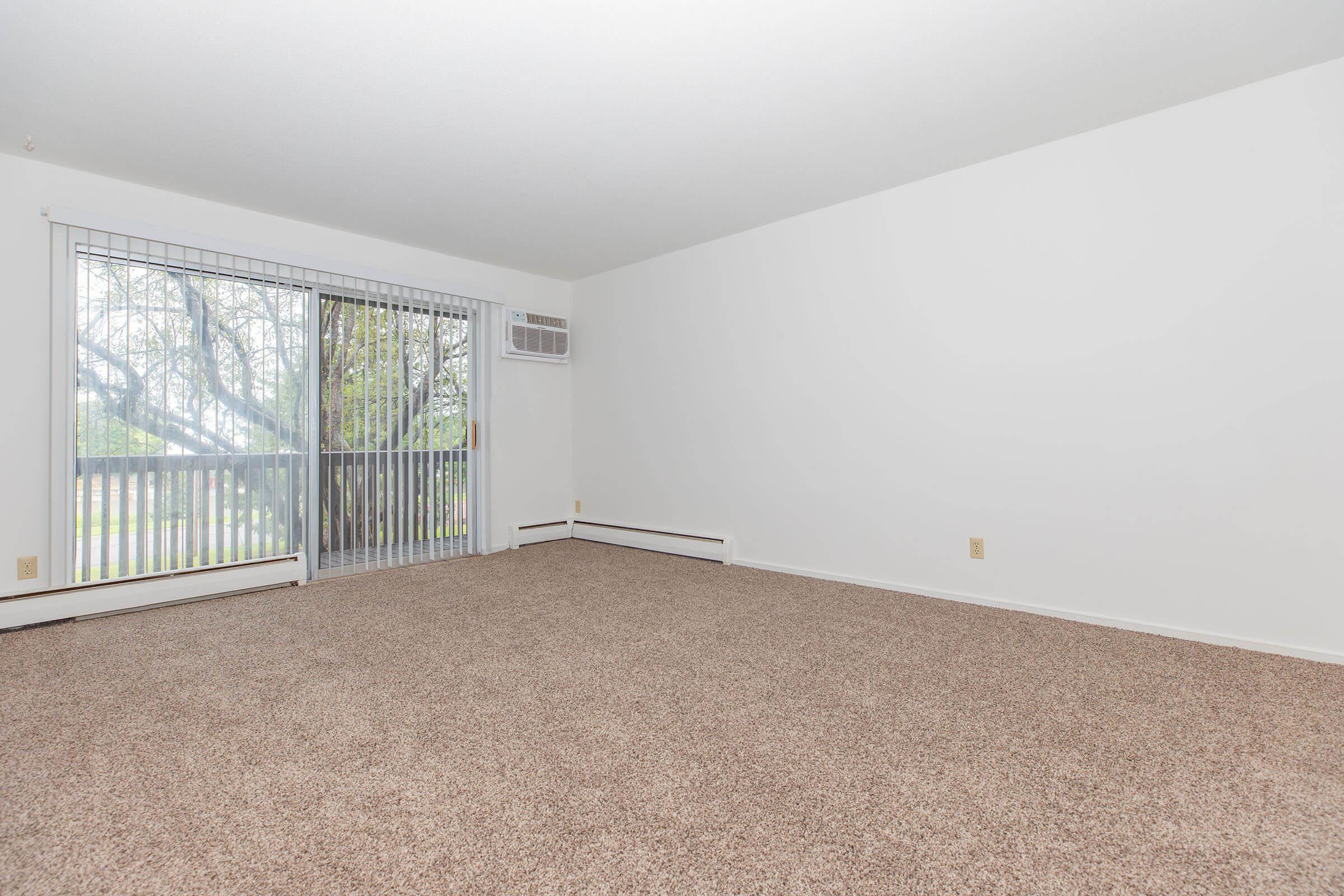 A spacious, empty living room with light beige carpet and white walls. Large sliding glass doors with vertical blinds allow natural light in, offering a view of greenery outside. An air conditioning unit is mounted in the wall. The room's simplicity suggests potential for personalization and decoration.