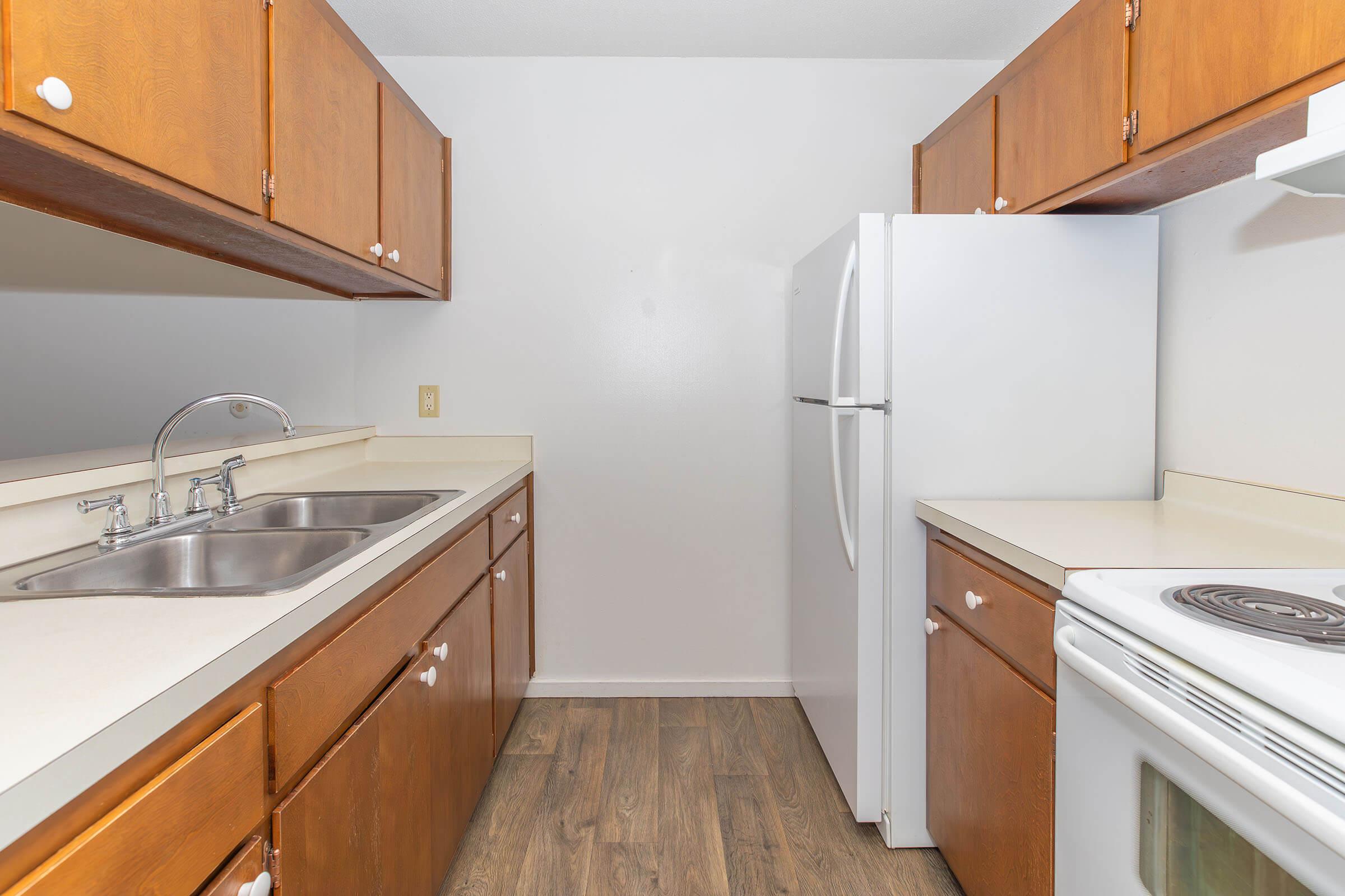 A small kitchen featuring wooden cabinets, a double sink, a white refrigerator, and a white stove. The countertops are light-colored, and the floor has a wood-like laminate finish. The walls are painted in a neutral tone, creating a bright and clean atmosphere.