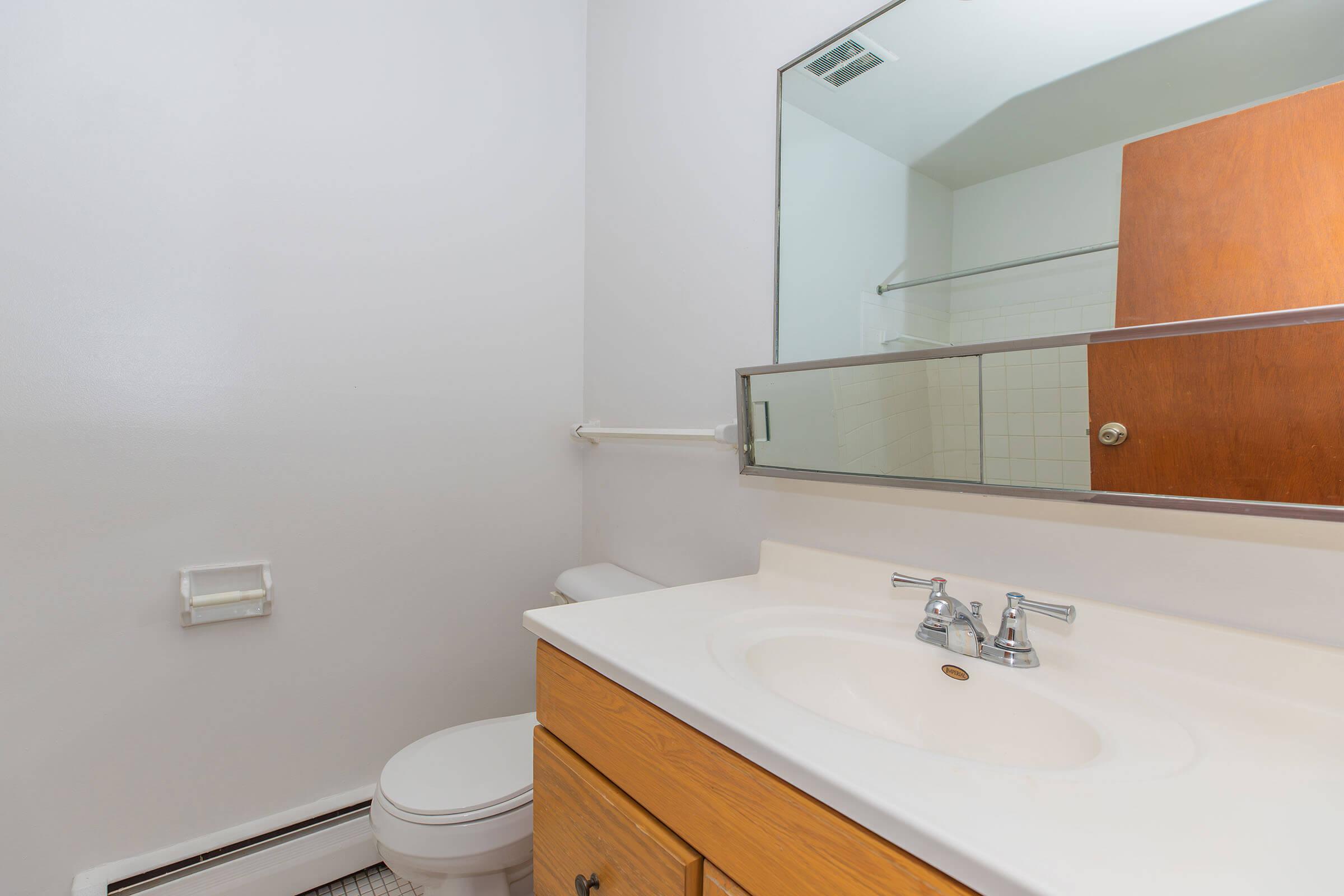 A clean, well-lit bathroom featuring a white toilet, a sink with two faucets, and a wooden cabinet below. A large mirror above the sink reflects the space, while a shower area is visible in the background. The walls are painted white, contributing to a bright and simple aesthetic.